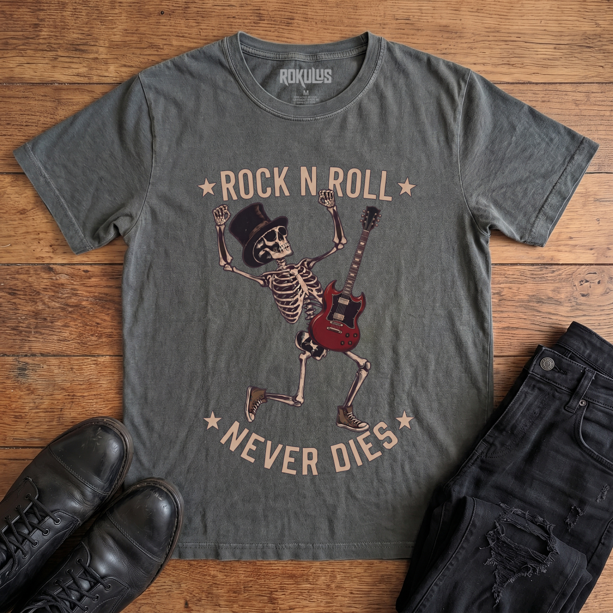 Washed charcoal Rokulus Rock N Roll Never Dies vintage graphic tee laid flat on wood with black boots and jeans, front print showing a dancing top-hat skeleton with a red electric guitar and fists raised under arched "Rock N Roll Never Dies" lettering