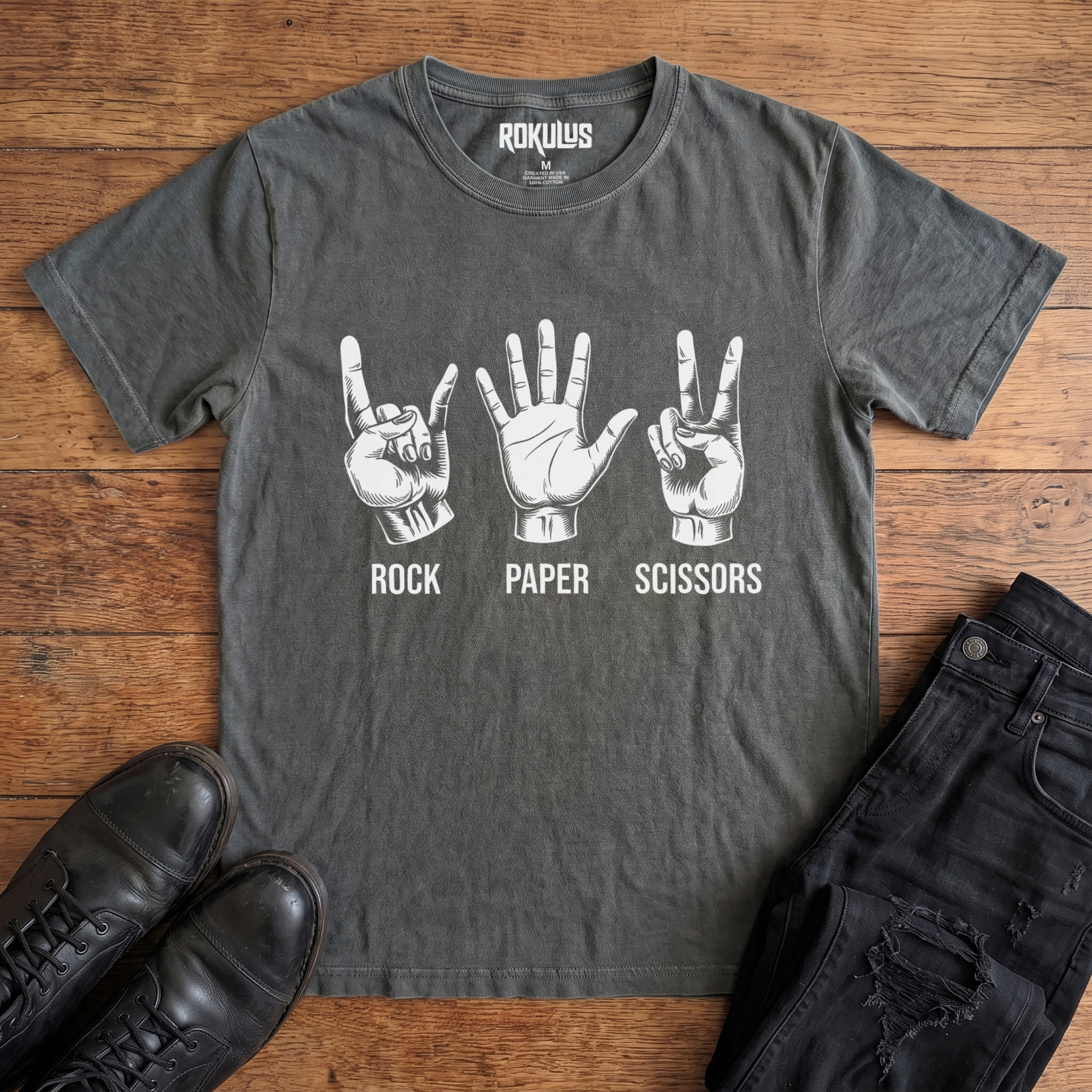 Washed charcoal Rokulus Rock Paper Scissors vintage graphic tee laid flat on wood with black boots and jeans, front print showing three hands in vintage engraving, devil horns for rock, flat palm for paper, peace sign for scissors