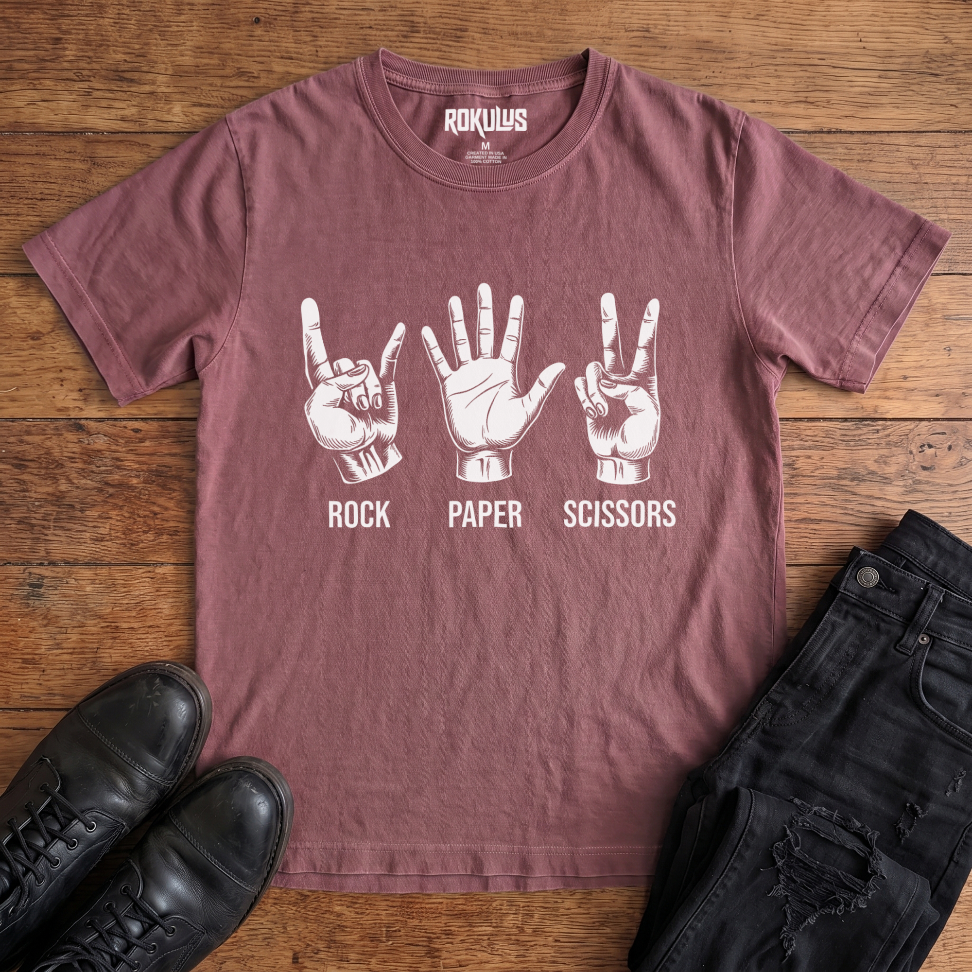 Washed maroon Rokulus Rock Paper Scissors vintage graphic tee laid flat on wood with black boots and jeans, front print showing three hands in vintage engraving, devil horns for rock, flat palm for paper, peace sign for scissors