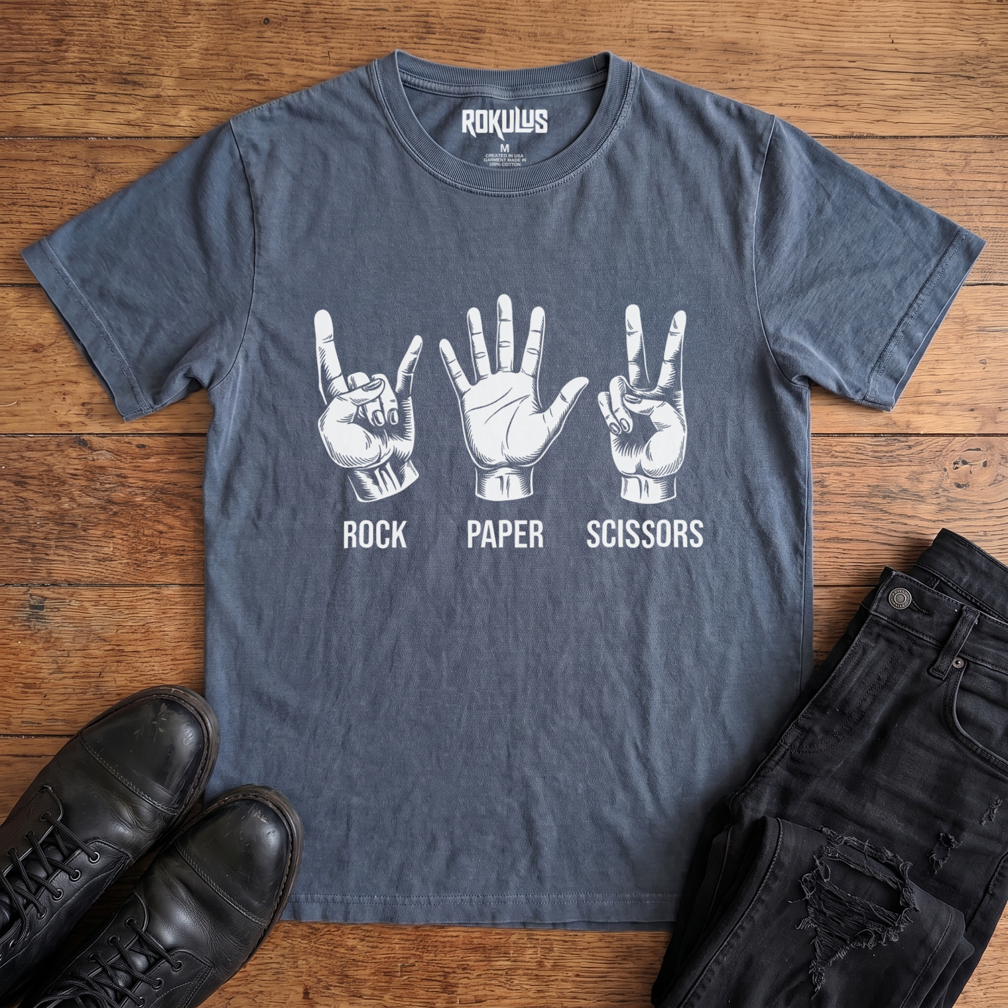 Washed navy blue Rokulus Rock Paper Scissors vintage graphic tee laid flat on wood with black boots and jeans, front print showing three hands in vintage engraving, devil horns for rock, flat palm for paper, peace sign for scissors