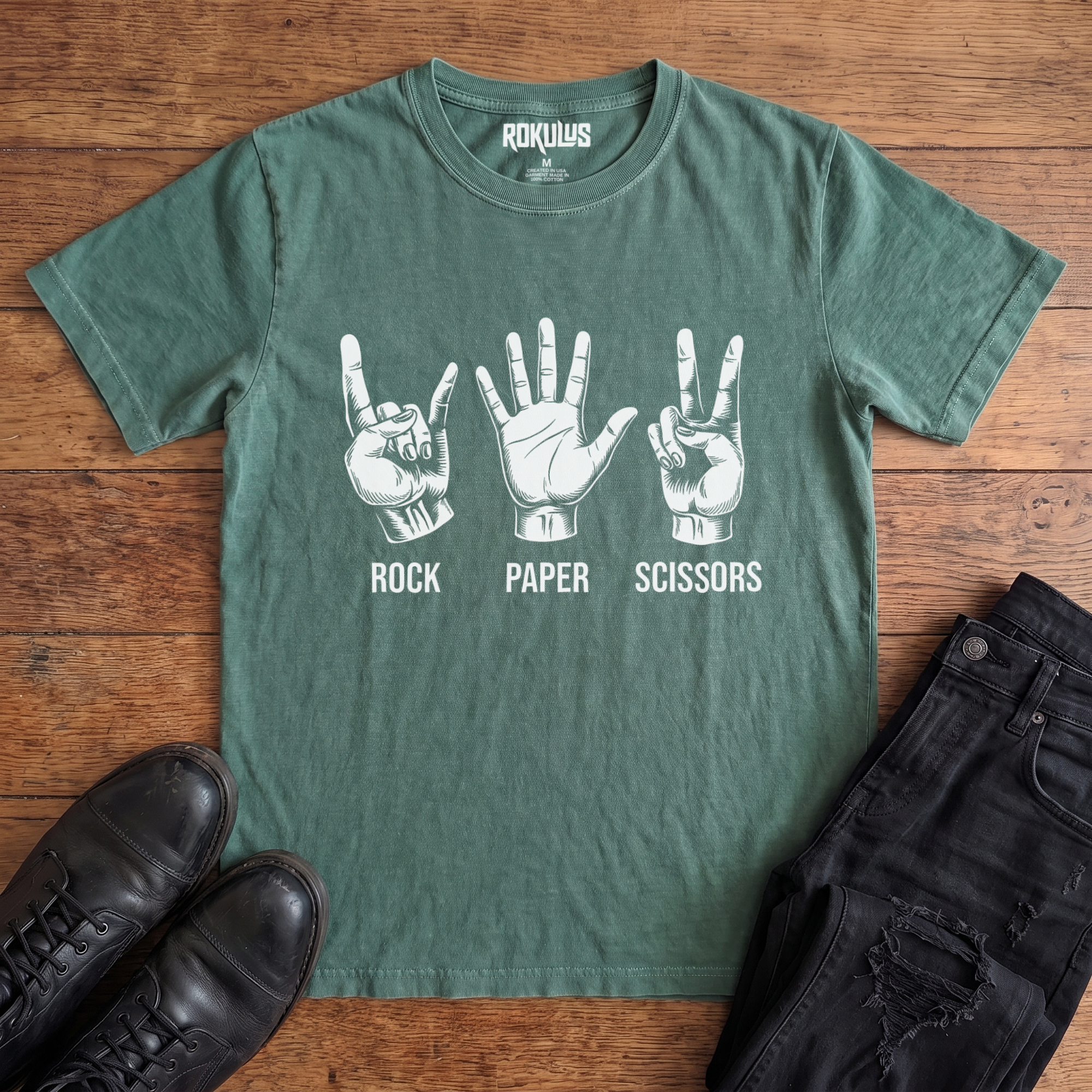Washed pine Rokulus Rock Paper Scissors vintage graphic tee laid flat on wood with black boots and jeans, front print showing three hands in vintage engraving, devil horns for rock, flat palm for paper, peace sign for scissors