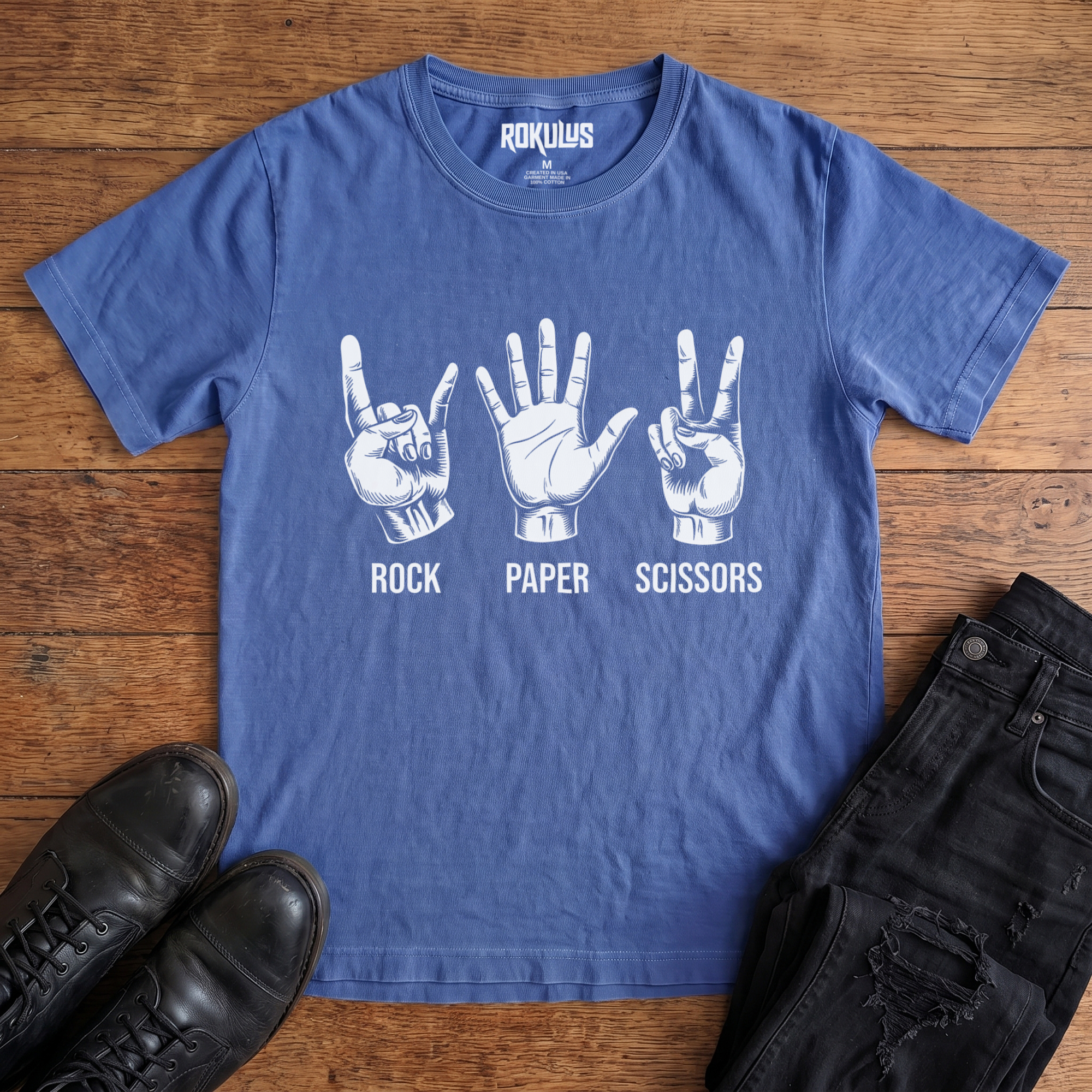 Washed royal blue Rokulus Rock Paper Scissors vintage graphic tee laid flat on wood with black boots and jeans, front print showing three hands in vintage engraving, devil horns for rock, flat palm for paper, peace sign for scissors