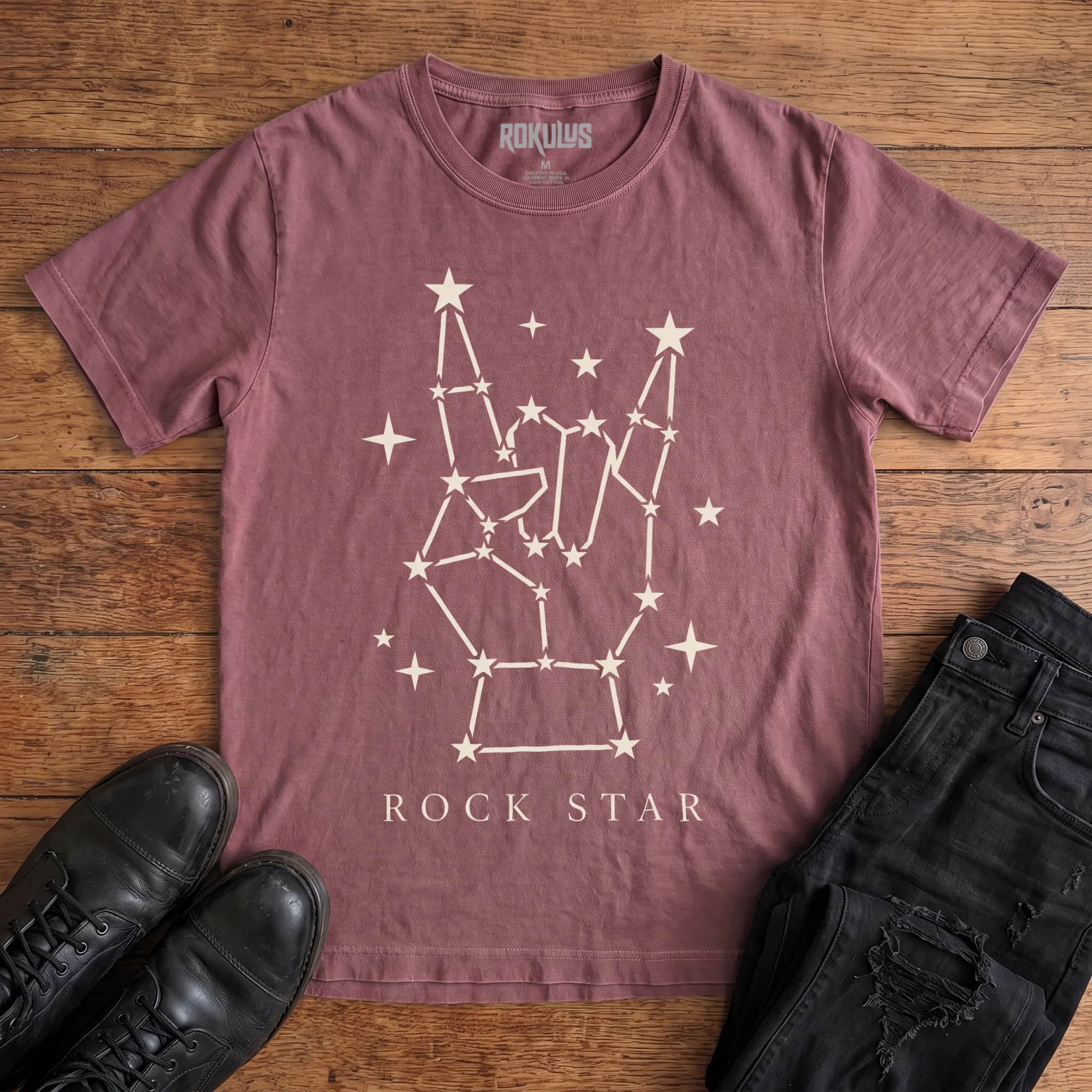 Washed maroon Rokulus Rock Star Constellation vintage graphic tee laid flat on wood with black boots and jeans, front print showing a rock horns hand formed from a star constellation above “Rock Star” lettering in cream