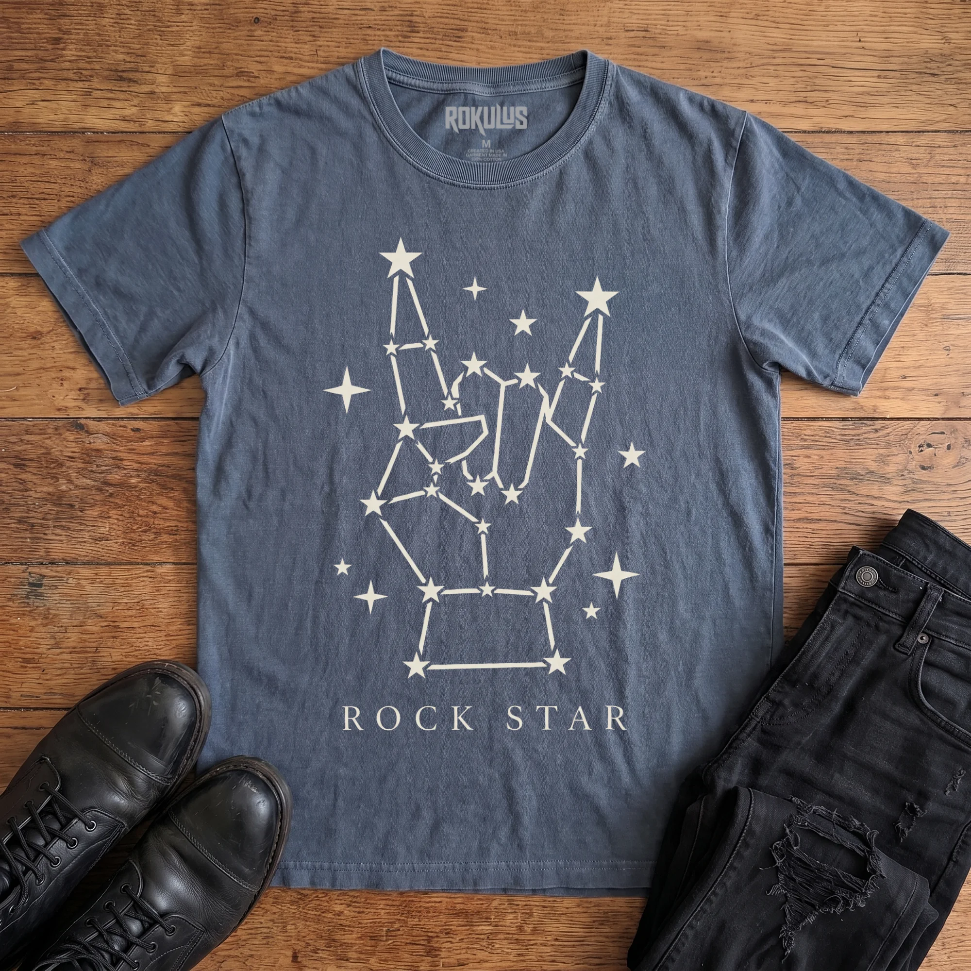 Washed navy Rokulus Rock Star Constellation vintage graphic tee laid flat on wood with black boots and jeans, front print showing a rock horns hand formed from a star constellation above “Rock Star” lettering in cream
