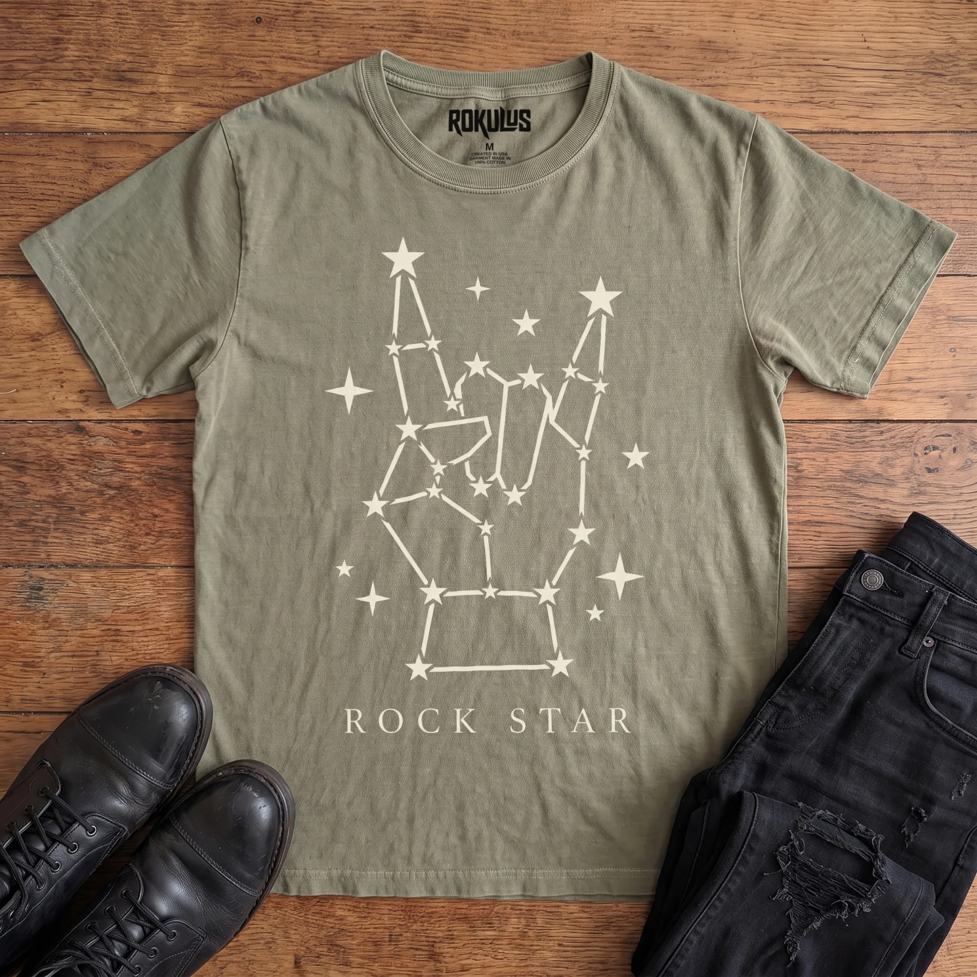 Washed olive Rokulus Rock Star Constellation vintage graphic tee laid flat on wood with black boots and jeans, front print showing a rock horns hand formed from a star constellation above “Rock Star” lettering in cream