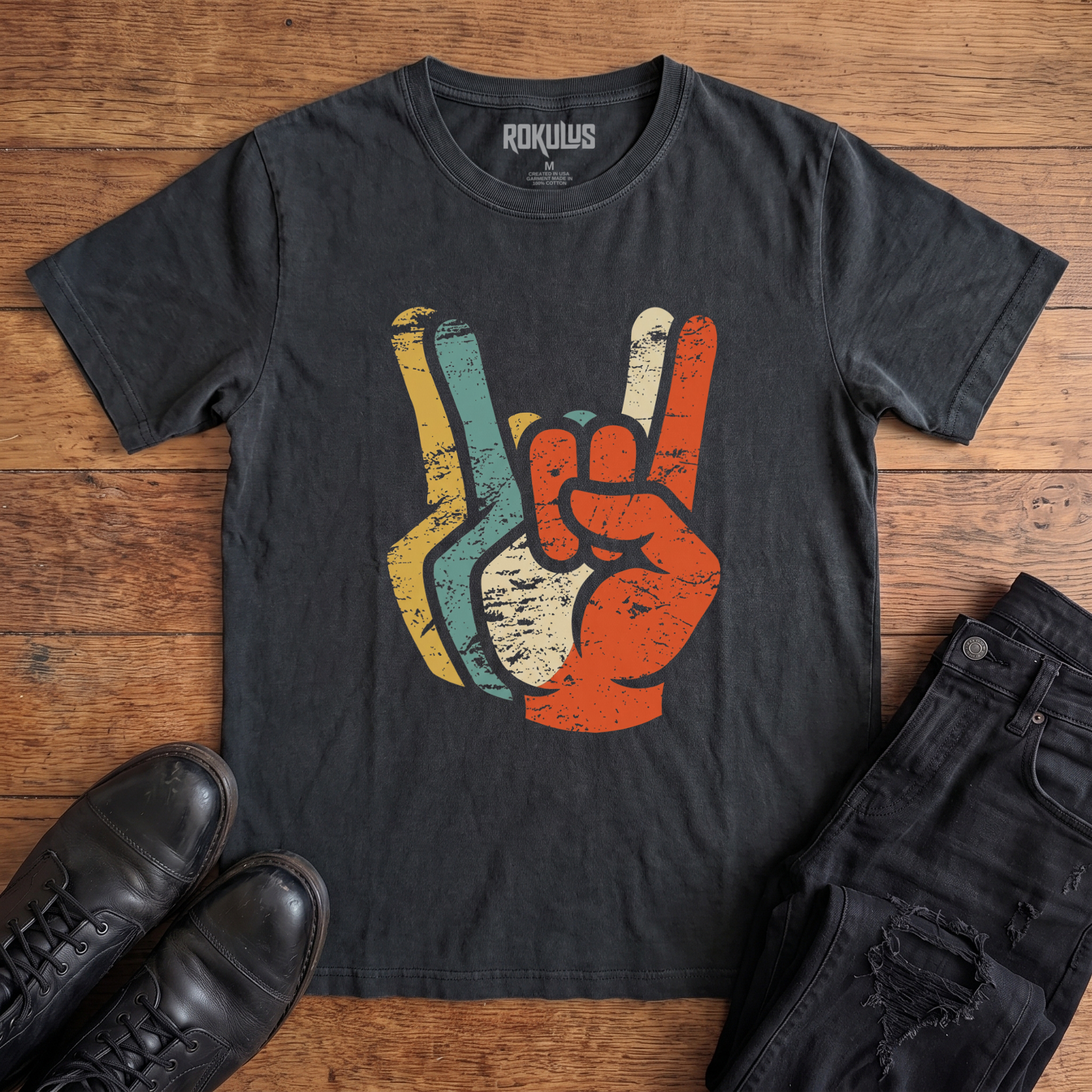Washed black Rokulus Retro Rocker Hands vintage graphic tee laid flat on wood with black boots and jeans, front print showing a devil-horns rock salute layered in distressed retro mustard, teal, cream, and burnt orange tones