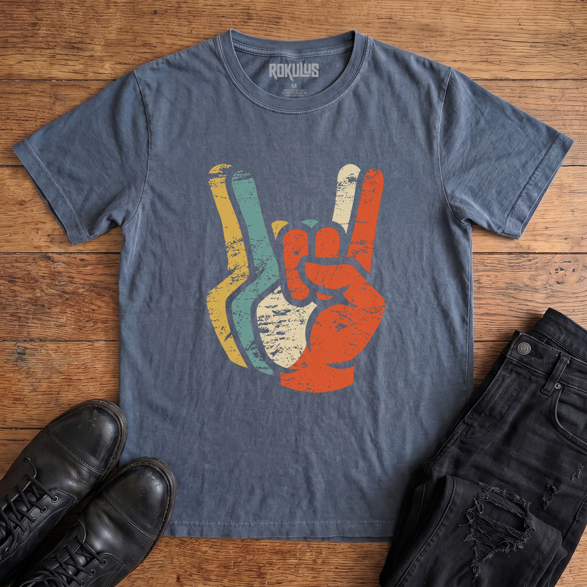 Washed navy Rokulus Retro Rocker Hands vintage graphic tee laid flat on wood with black boots and jeans, front print showing a devil-horns rock salute layered in distressed retro mustard, teal, cream, and burnt orange tones