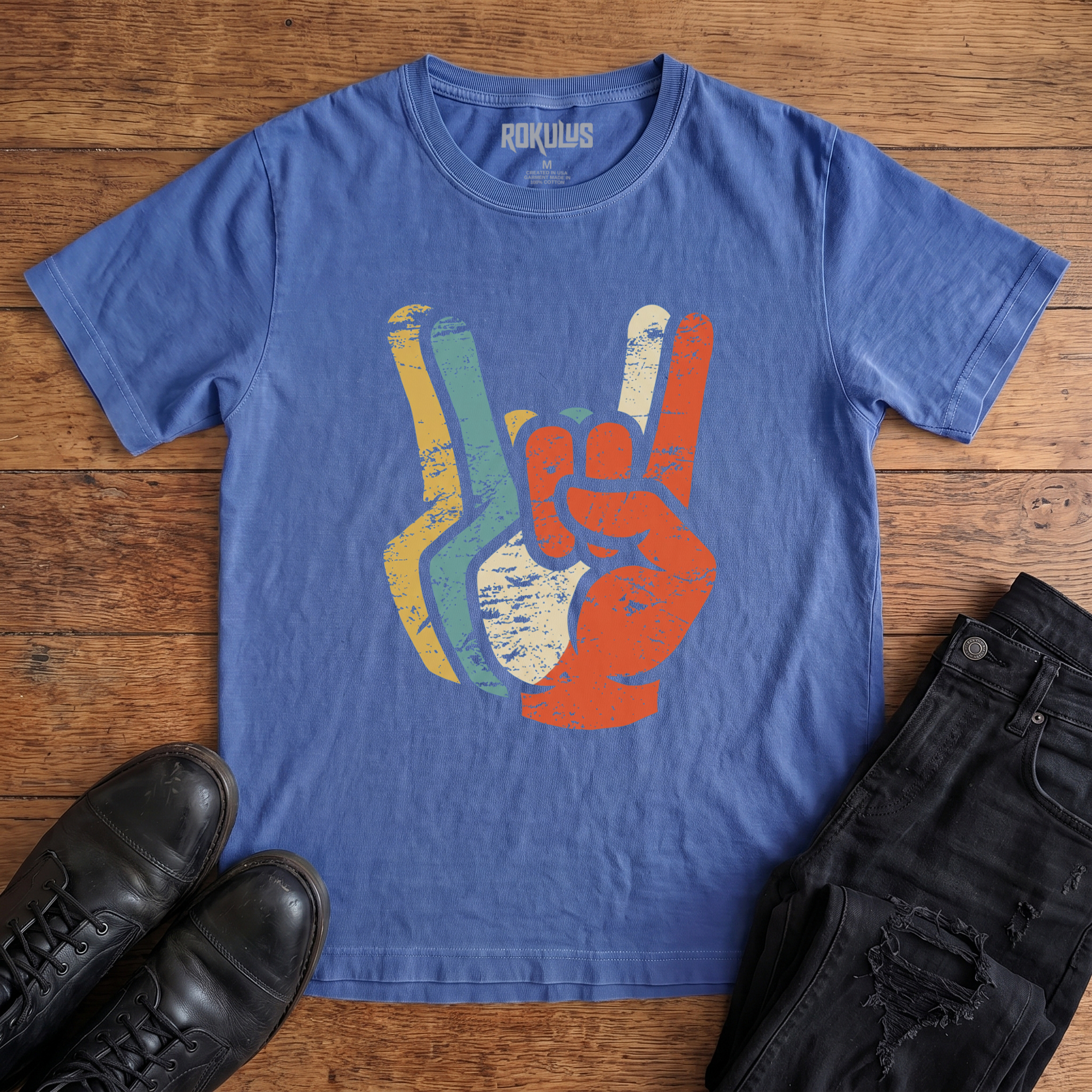 Washed royal blue Rokulus Retro Rocker Hands vintage graphic tee laid flat on wood with black boots and jeans, front print showing a devil-horns rock salute layered in distressed retro mustard, teal, cream, and burnt orange tones