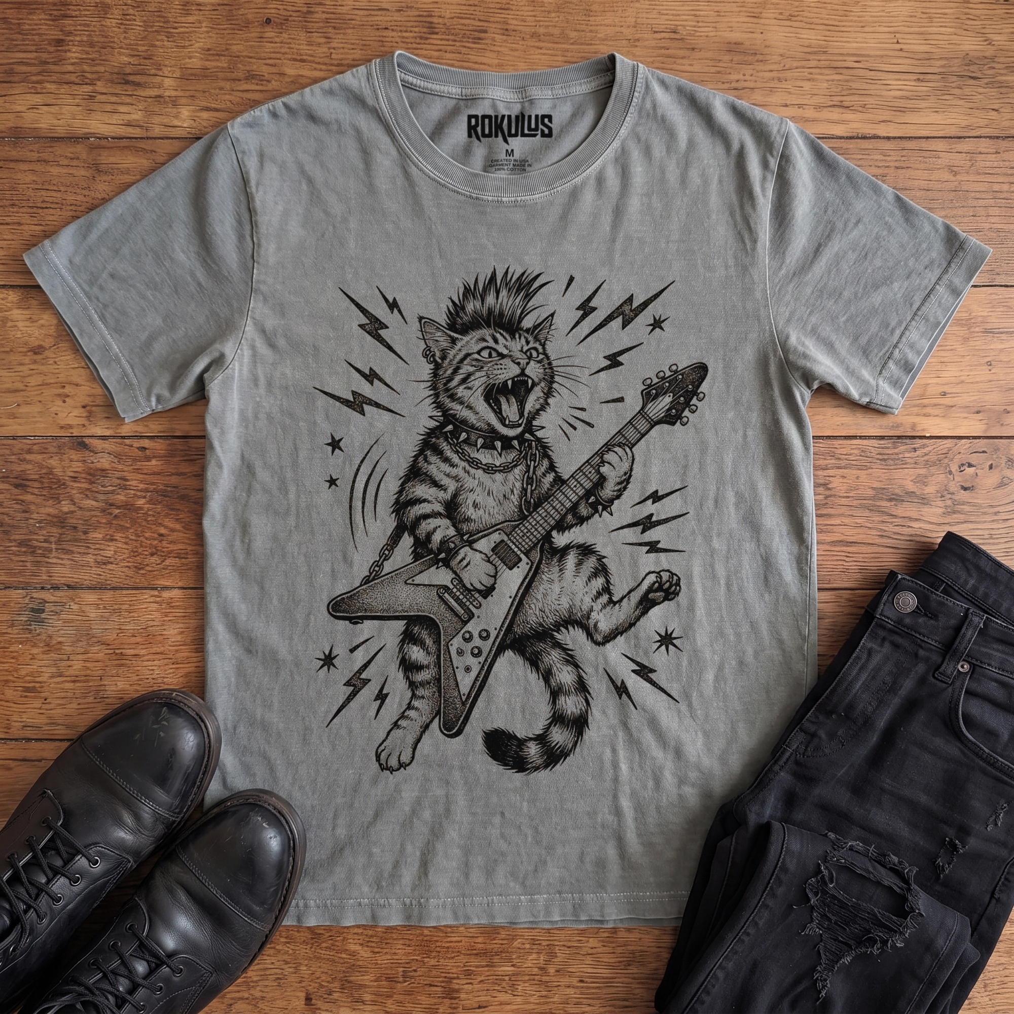 Washed grey Rokulus Rocking Cat vintage graphic tee laid flat on wood with black boots and jeans, front print showing a mohawked cat shredding a Flying V guitar in bold black linework with lightning bolts and stars