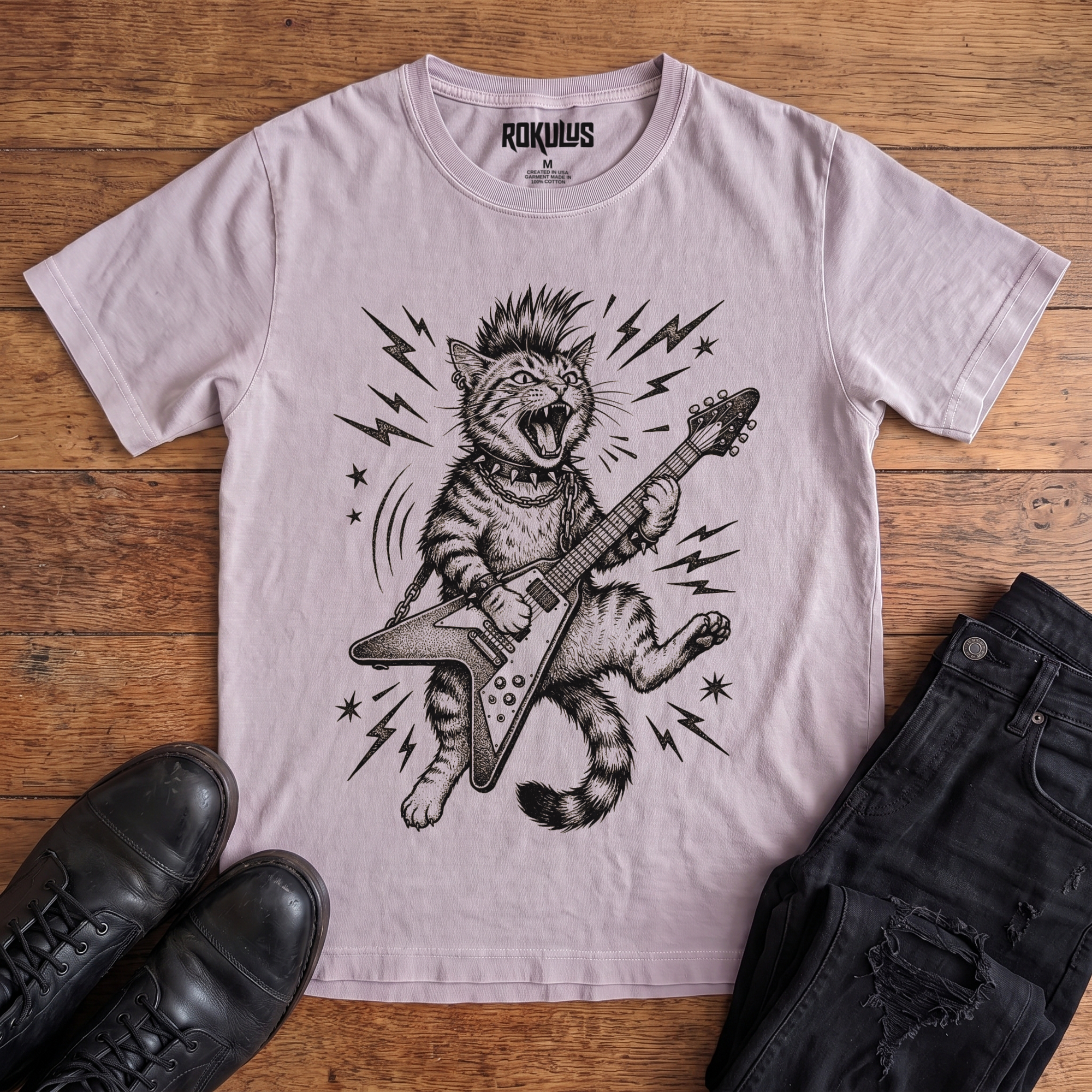 Washed lavender Rokulus Rocking Cat vintage graphic tee laid flat on wood with black boots and jeans, front print showing a mohawked cat shredding a Flying V guitar in bold black linework with lightning bolts and stars
