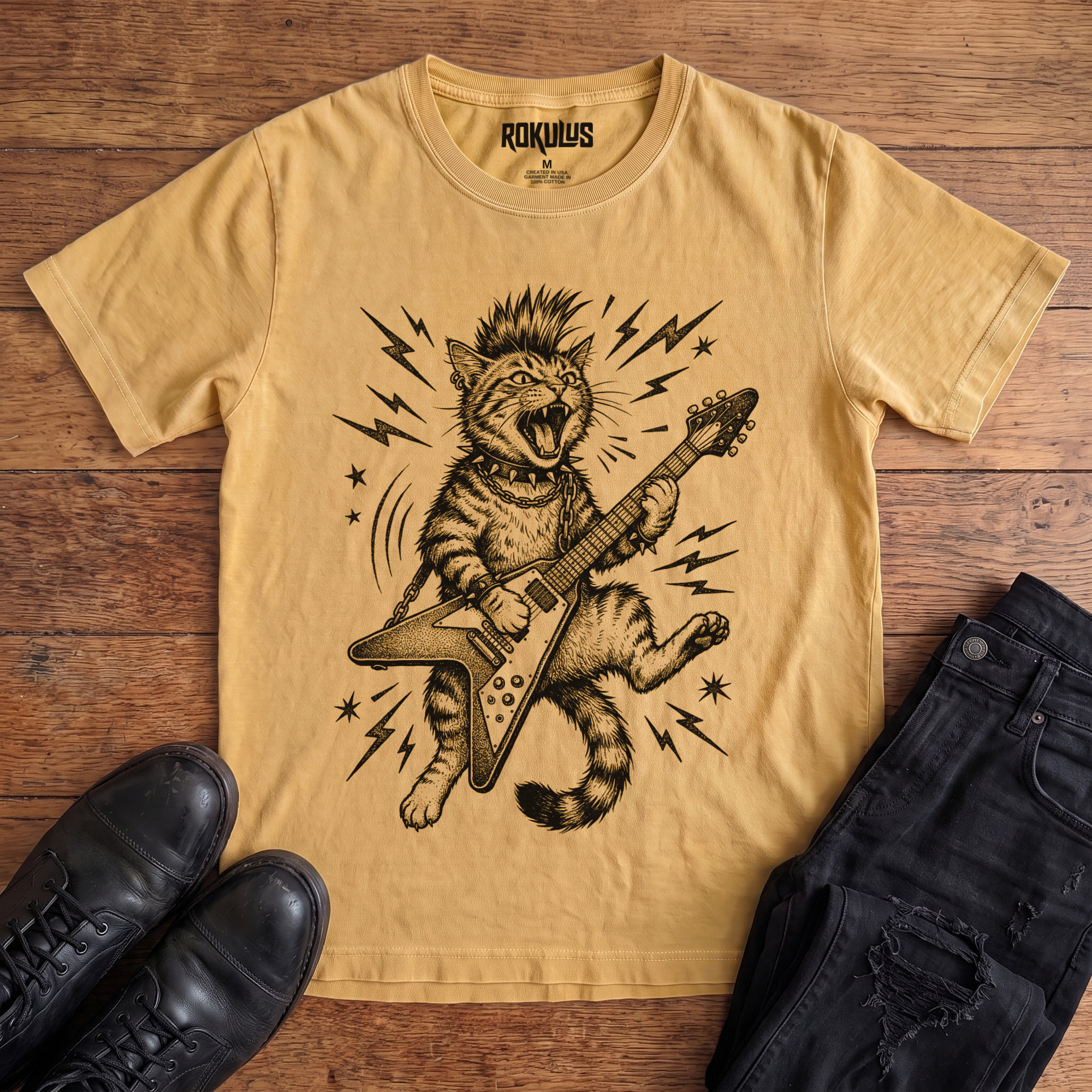 Washed mustard yellow Rokulus Rocking Cat vintage graphic tee laid flat on wood with black boots and jeans, front print showing a mohawked cat shredding a Flying V guitar in bold black linework with lightning bolts and stars