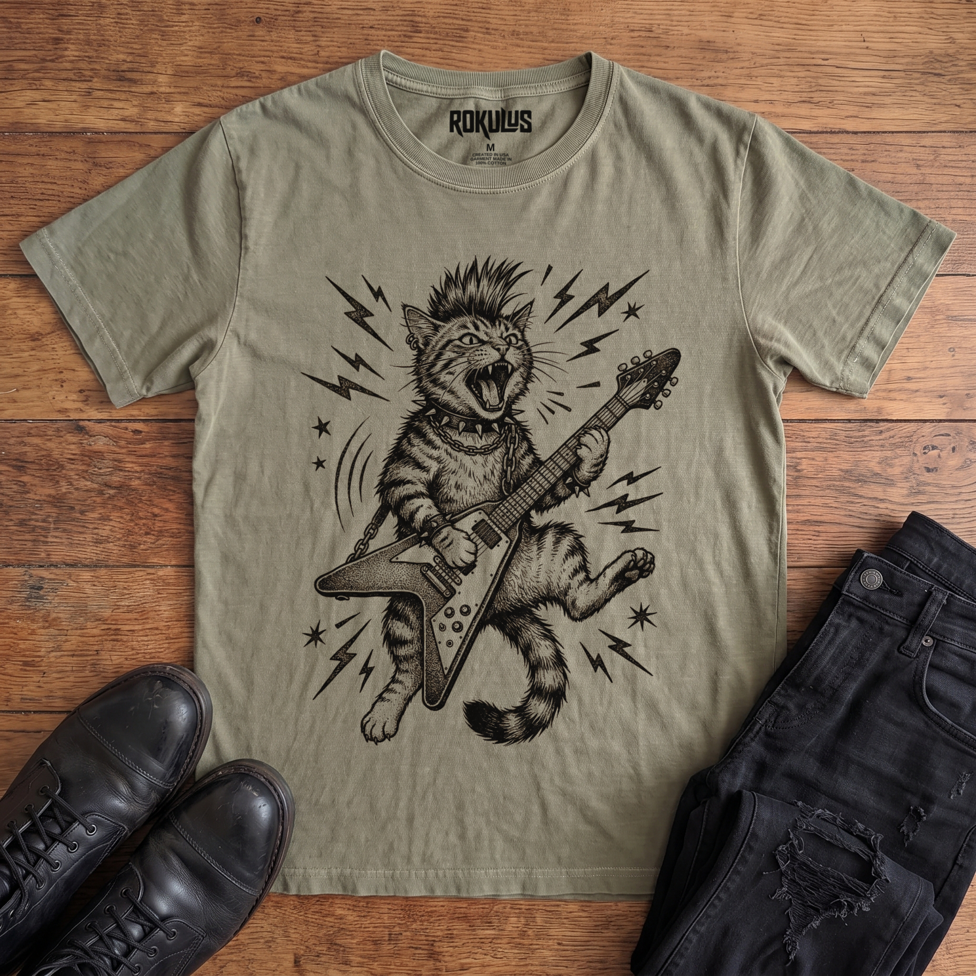 Washed olive Rokulus Rocking Cat vintage graphic tee laid flat on wood with black boots and jeans, front print showing a mohawked cat shredding a Flying V guitar in bold black linework with lightning bolts and stars