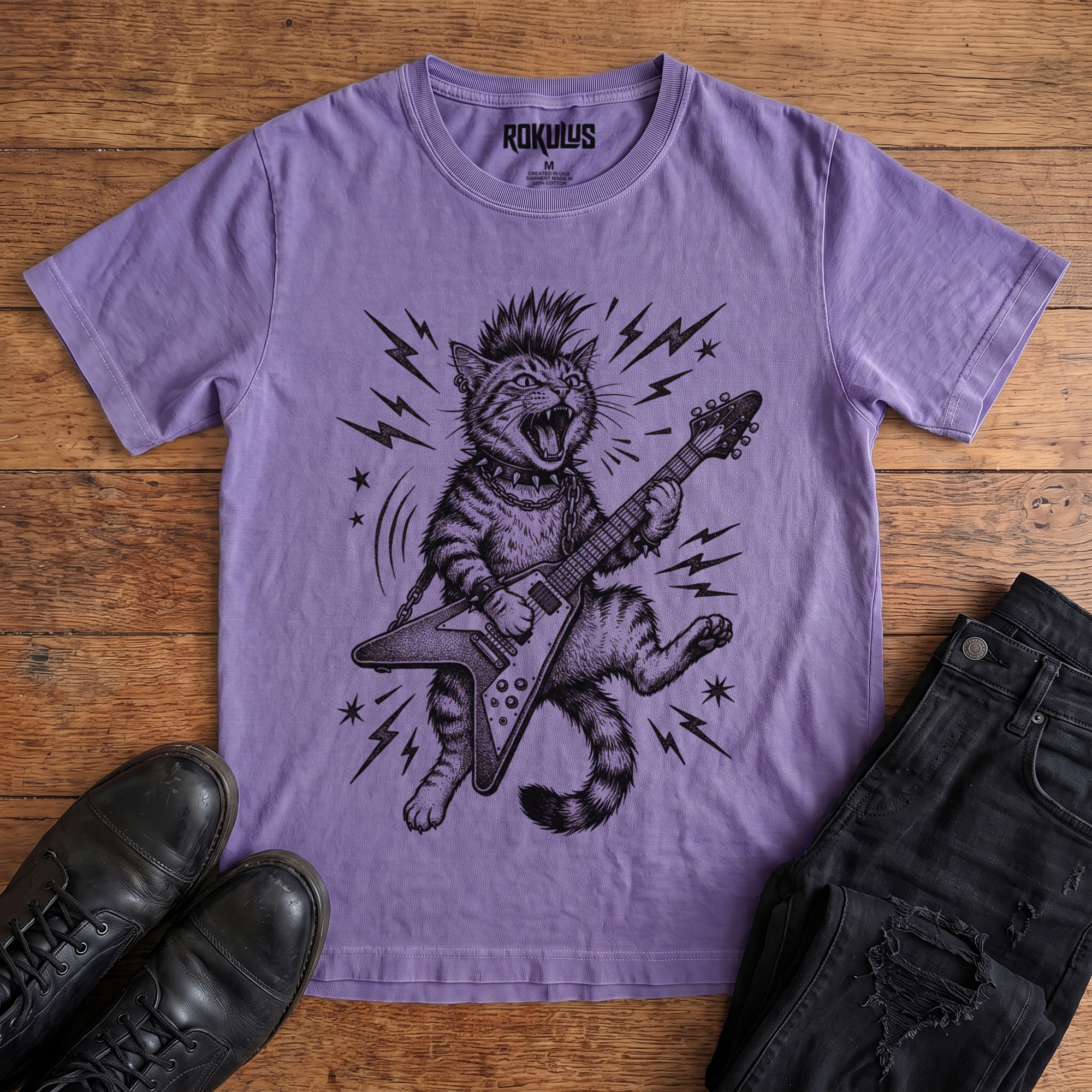 Washed purple Rokulus Rocking Cat vintage graphic tee laid flat on wood with black boots and jeans, front print showing a mohawked cat shredding a Flying V guitar in bold black linework with lightning bolts and stars