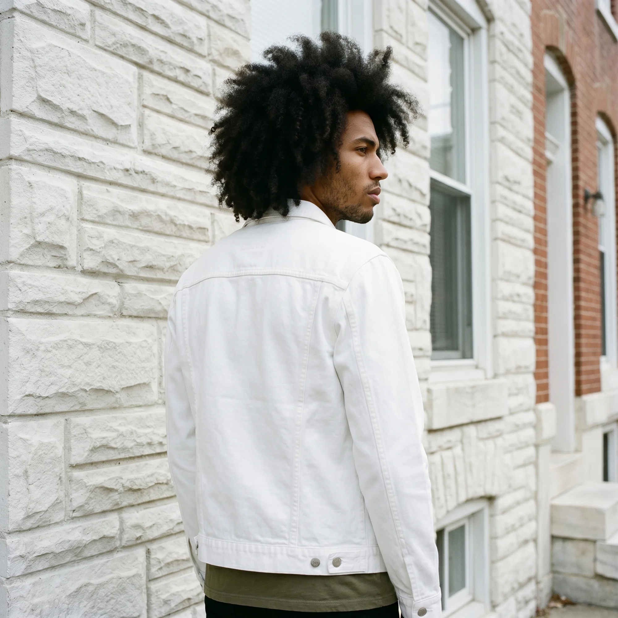 Man with an afro wearing a white denim jacket, back view, showing the trucker seam detailing against a stone row-house wall