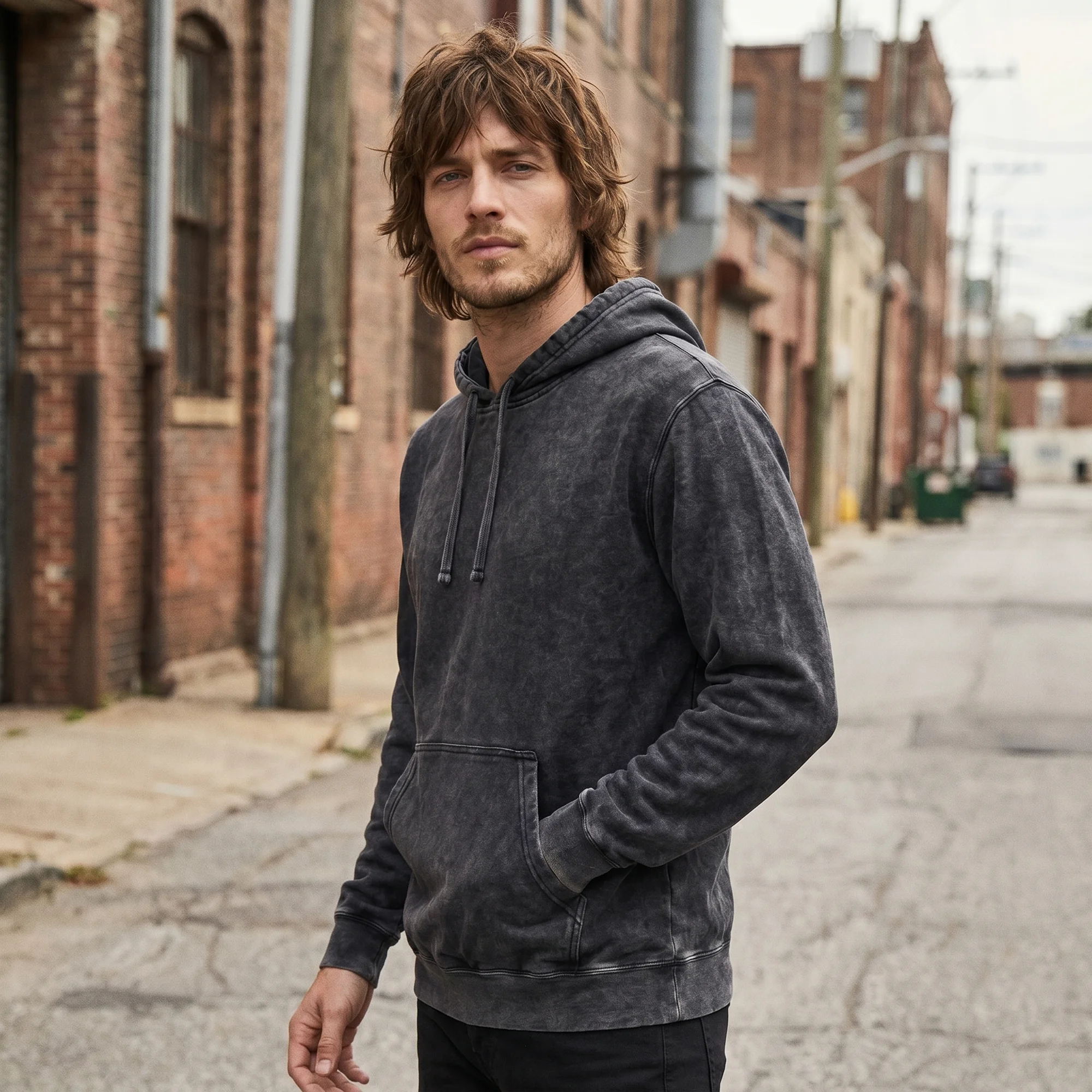 Man wearing a black mineral wash hoodie, side view, hood down, in a brick alley