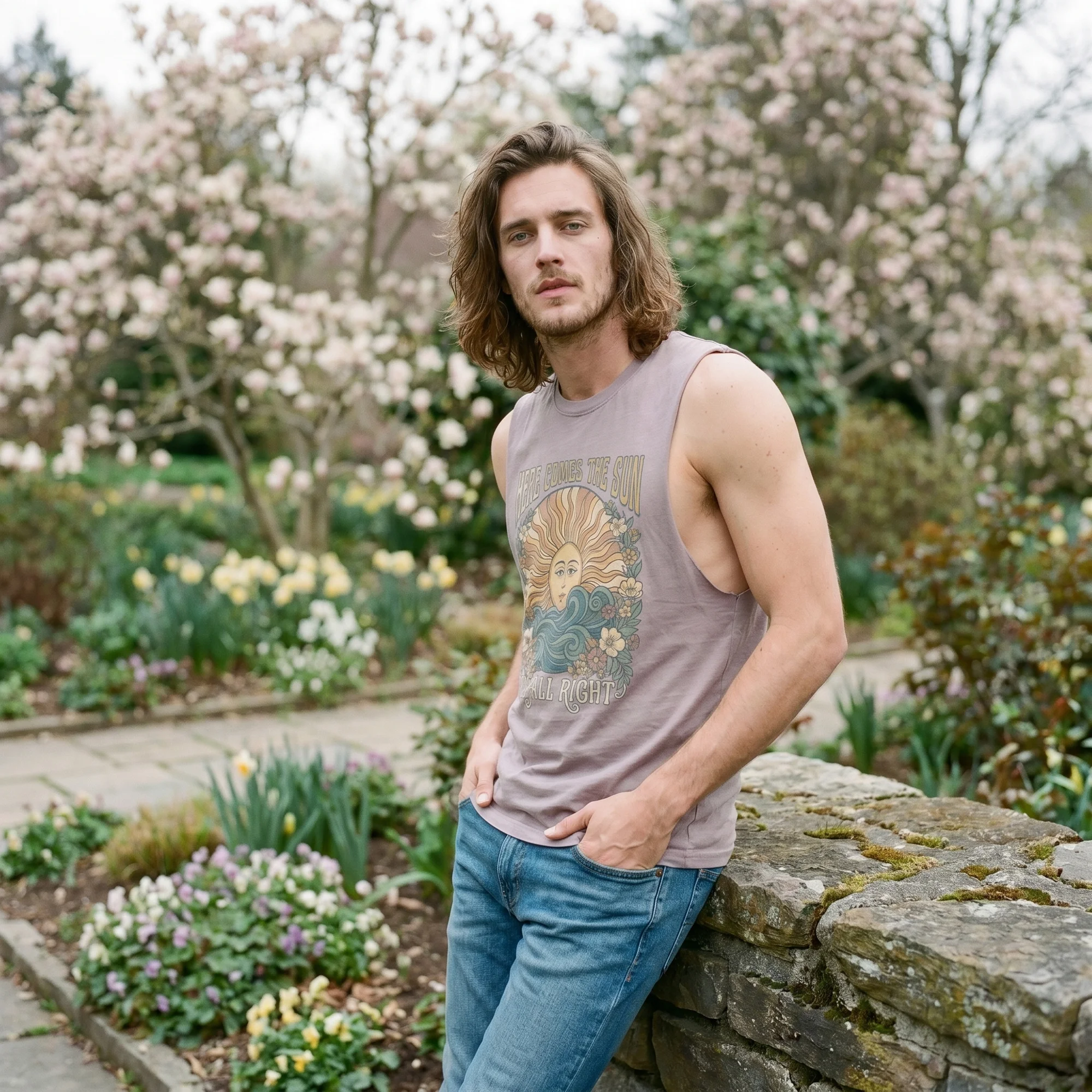 Man with long wavy hair leaning against a stone wall in a flower garden, wearing an orchid Here Comes the Sun tank top, side view