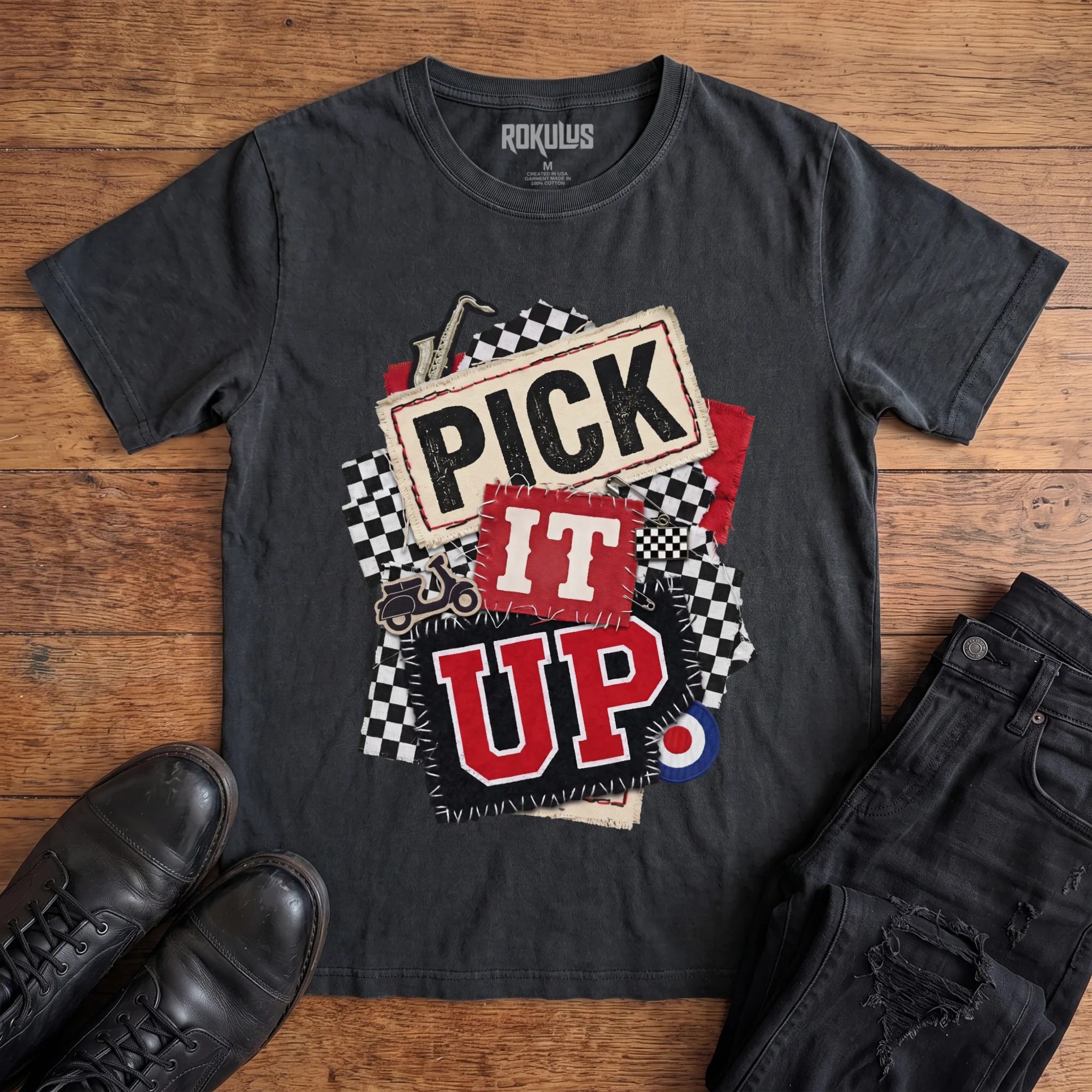 Washed black Rokulus Pick It Up Ska vintage graphic tee laid flat on wood with black boots and jeans, front print showing a 2-tone ska collage with checkerboard, saxophone, scooter, and mod target under stitched "Pick It Up" lettering