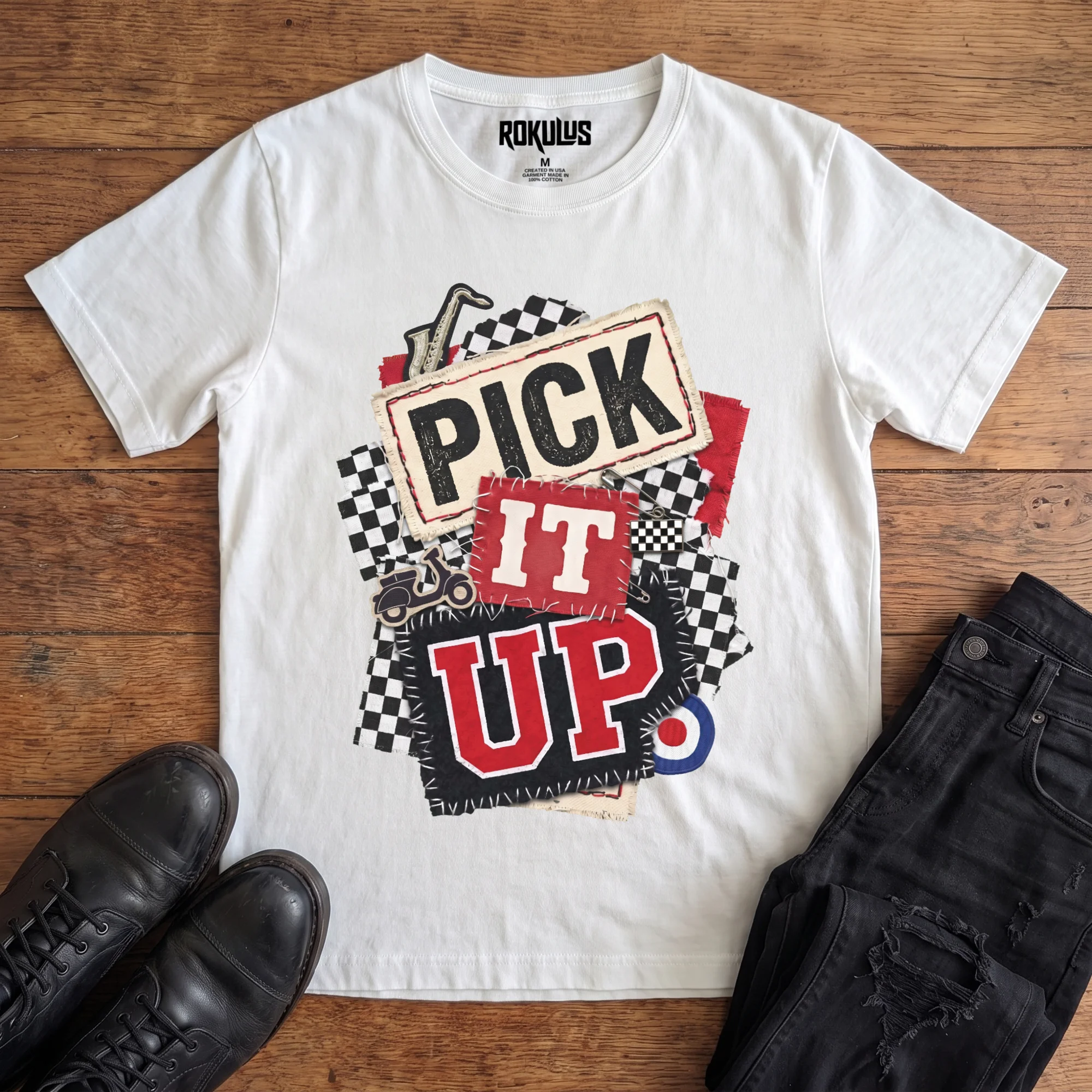 White Rokulus Pick It Up Ska vintage graphic tee laid flat on wood with black boots and jeans, front print showing a 2-tone ska collage with checkerboard, saxophone, scooter, and mod target under stitched "Pick It Up" lettering