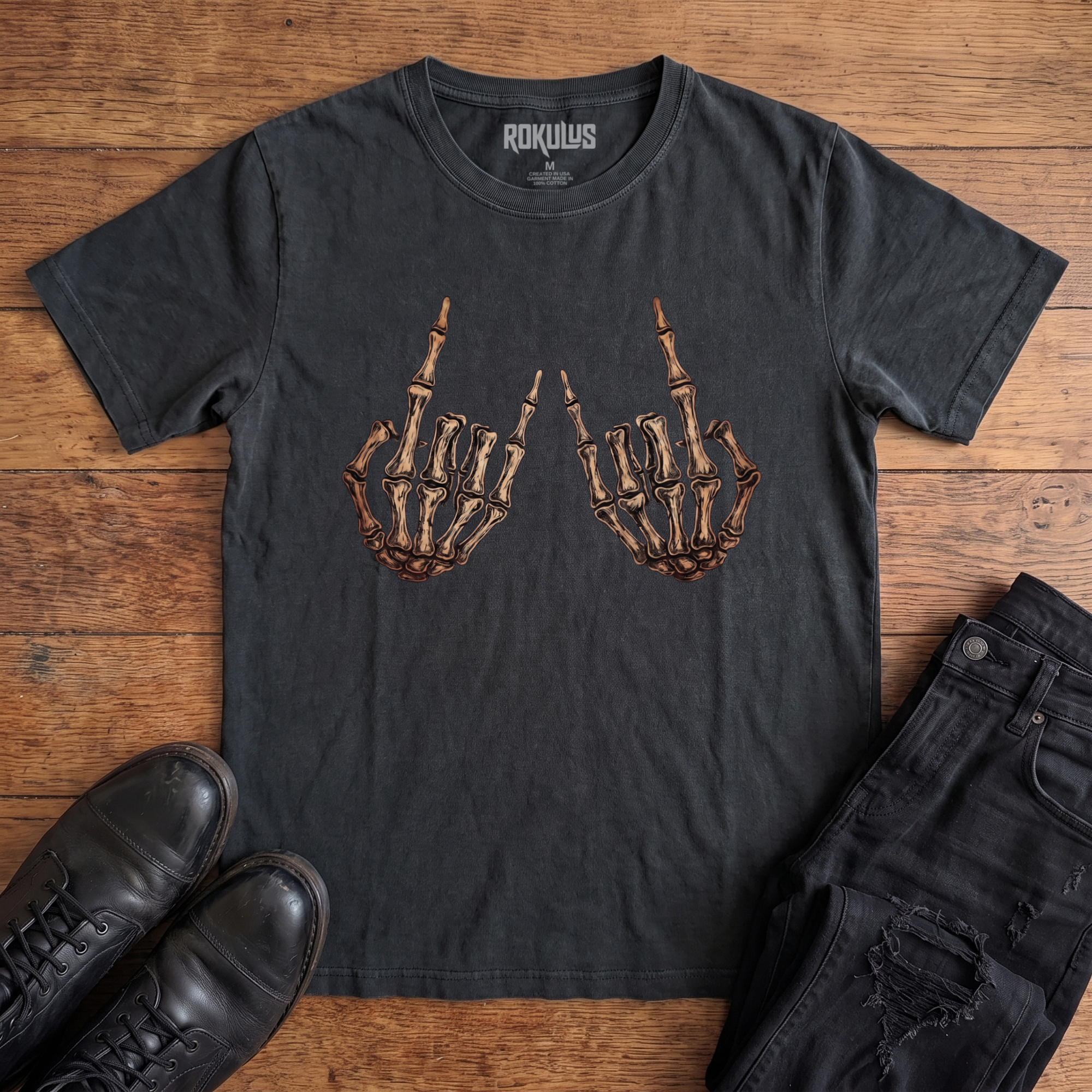Washed black Rokulus Skeleton Rocker Hands vintage graphic tee laid flat on wood with black boots and jeans, front print showing two skeletal hands thrown up in the rock and roll horns sign in detailed bone and cream tones