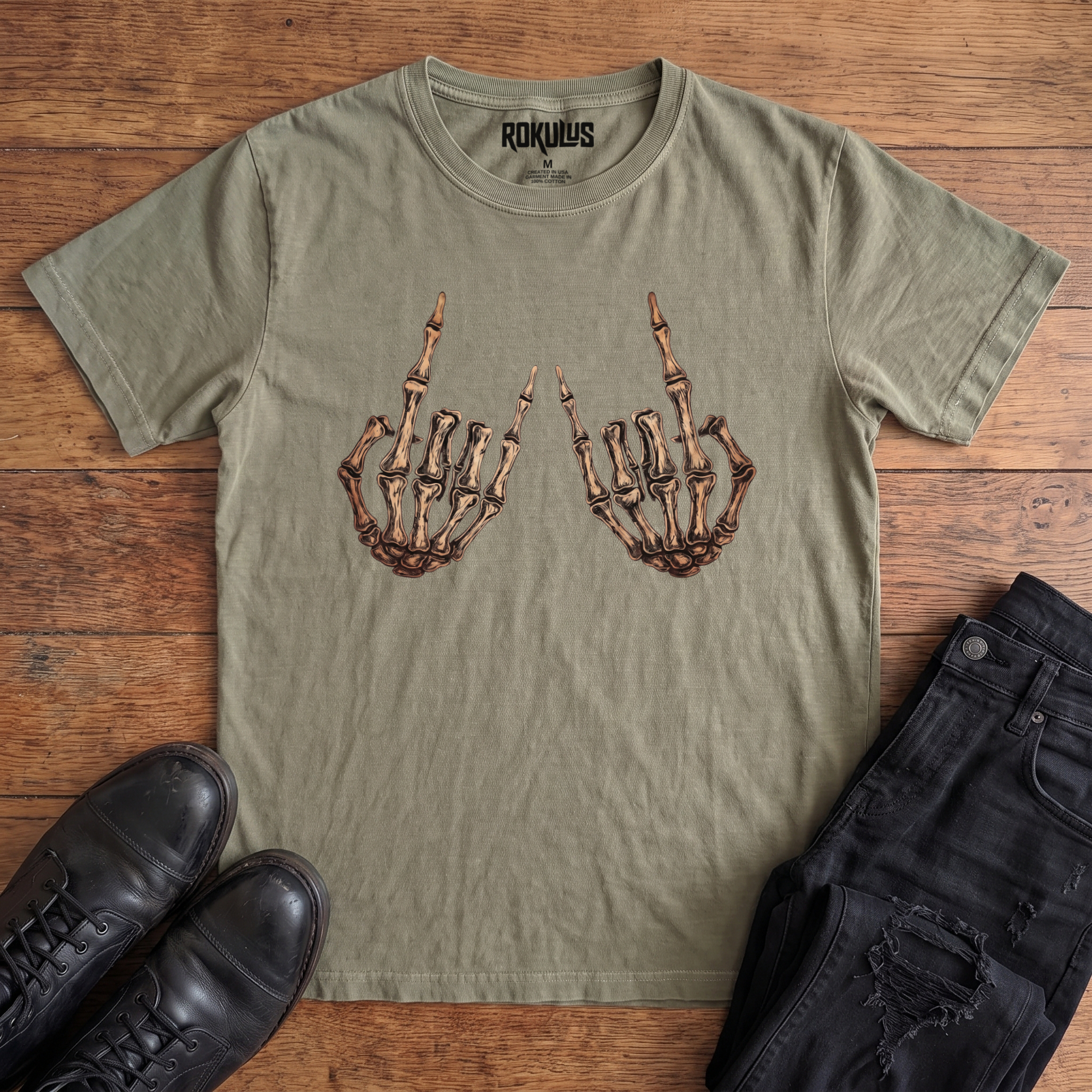Washed olive green Rokulus Skeleton Rocker Hands vintage graphic tee laid flat on wood with black boots and jeans, front print showing two skeletal hands thrown up in the rock and roll horns sign in detailed bone and cream tones