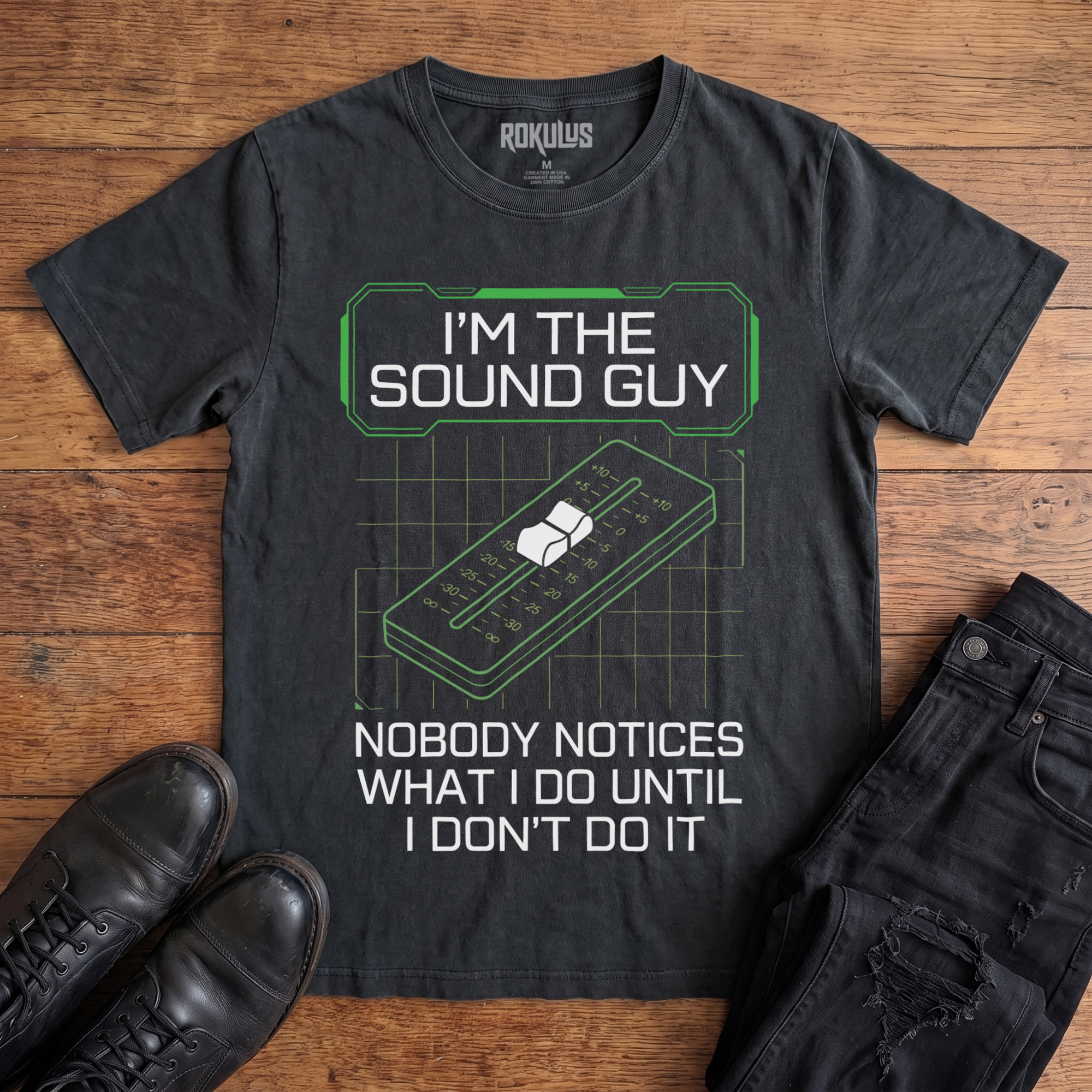 Washed black Rokulus Sound Guy vintage graphic tee laid flat on wood with black boots and jeans, front print reading "I'm the Sound Guy — Nobody Notices What I Do Until I Don't Do It" over a green mixing-console fader and grid