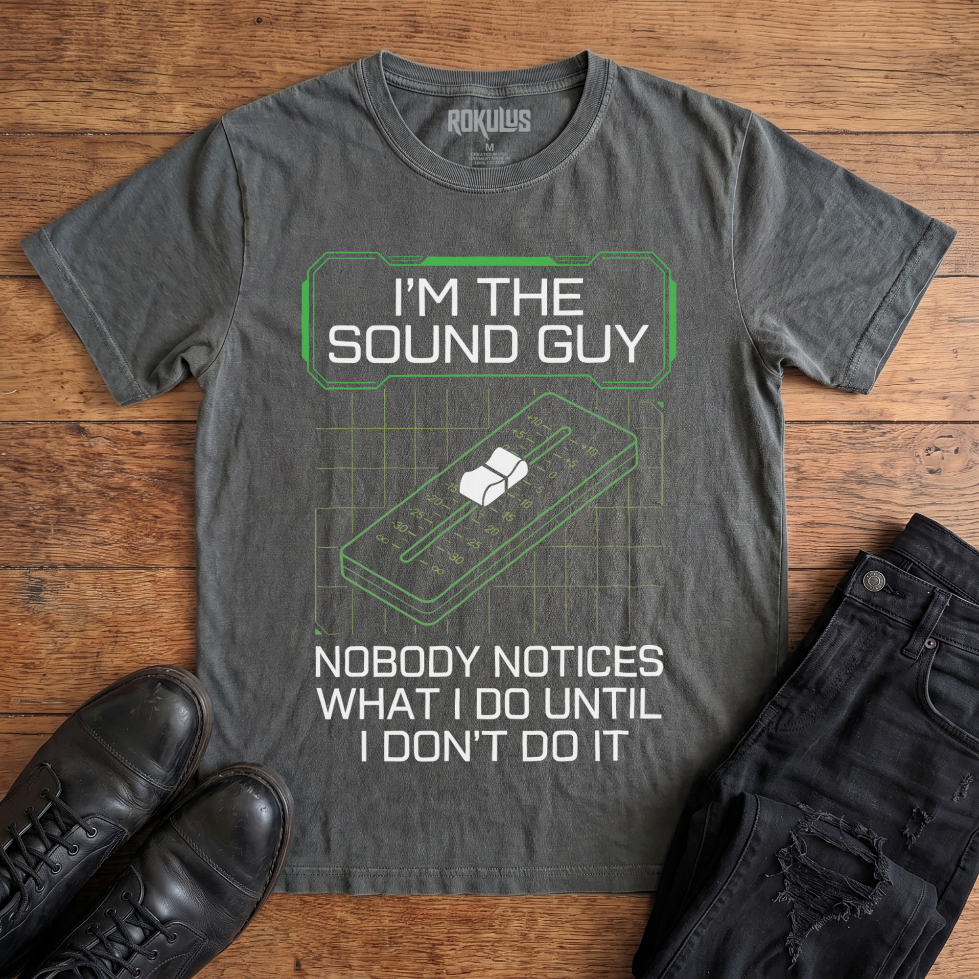 Washed charcoal Rokulus Sound Guy vintage graphic tee laid flat on wood with black boots and jeans, front print reading "I'm the Sound Guy — Nobody Notices What I Do Until I Don't Do It" over a green mixing-console fader and grid