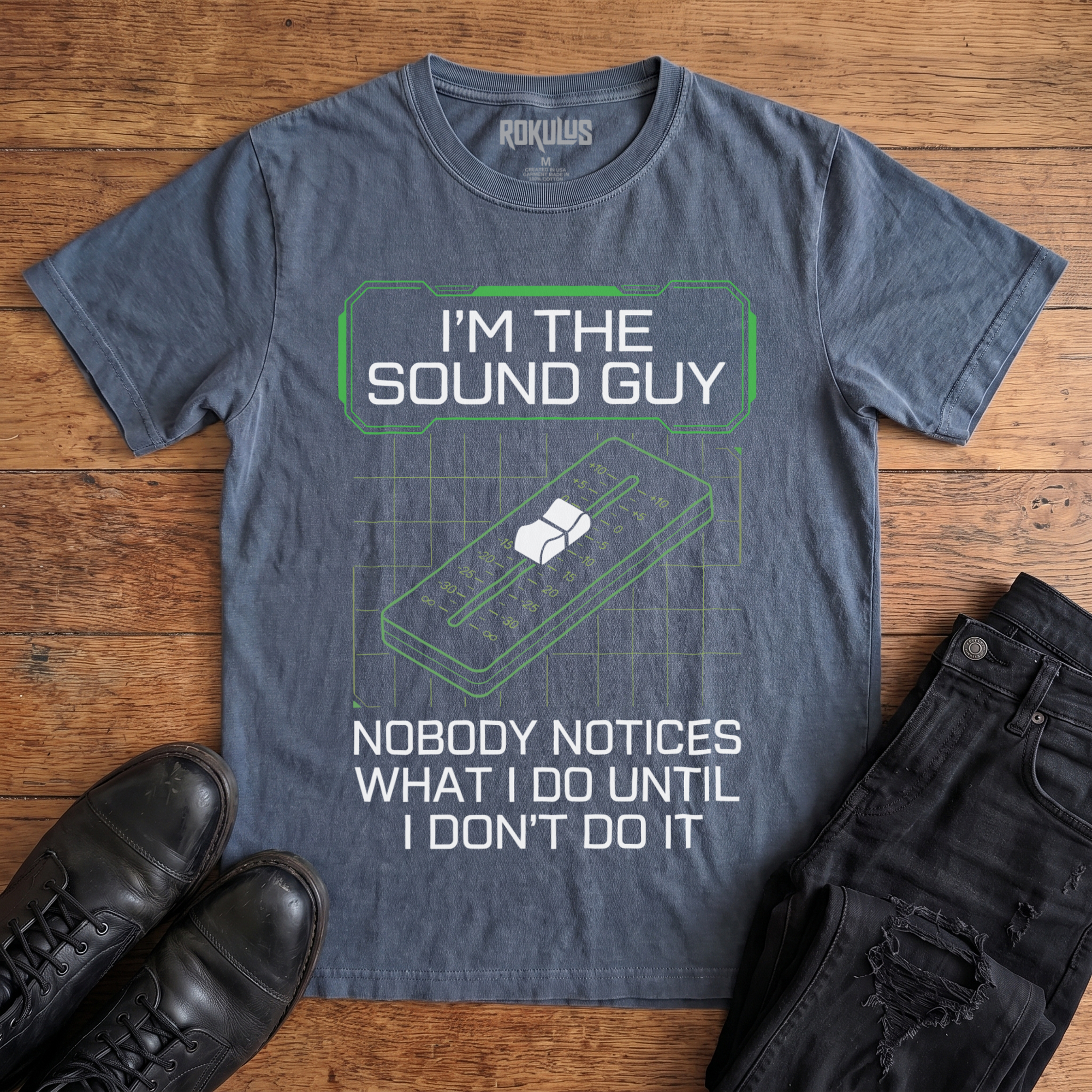 Washed navy Rokulus Sound Guy vintage graphic tee laid flat on wood with black boots and jeans, front print reading "I'm the Sound Guy — Nobody Notices What I Do Until I Don't Do It" over a green mixing-console fader and grid