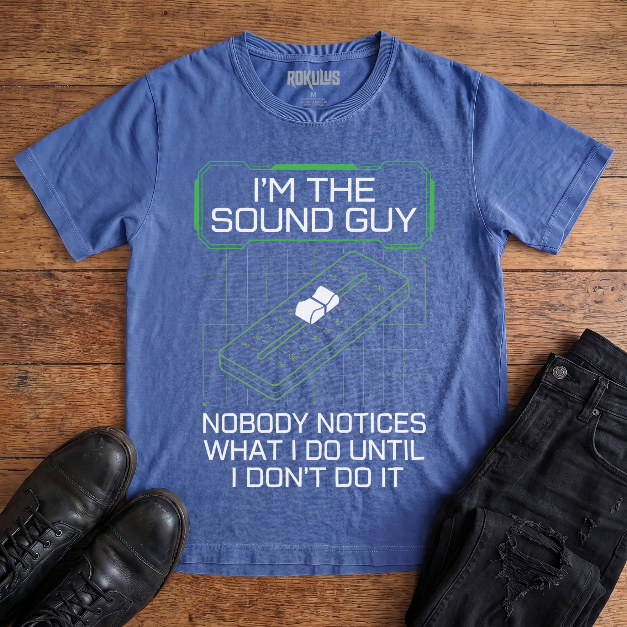 Washed royal blue Rokulus Sound Guy vintage graphic tee laid flat on wood with black boots and jeans, front print reading "I'm the Sound Guy — Nobody Notices What I Do Until I Don't Do It" over a green mixing-console fader and grid