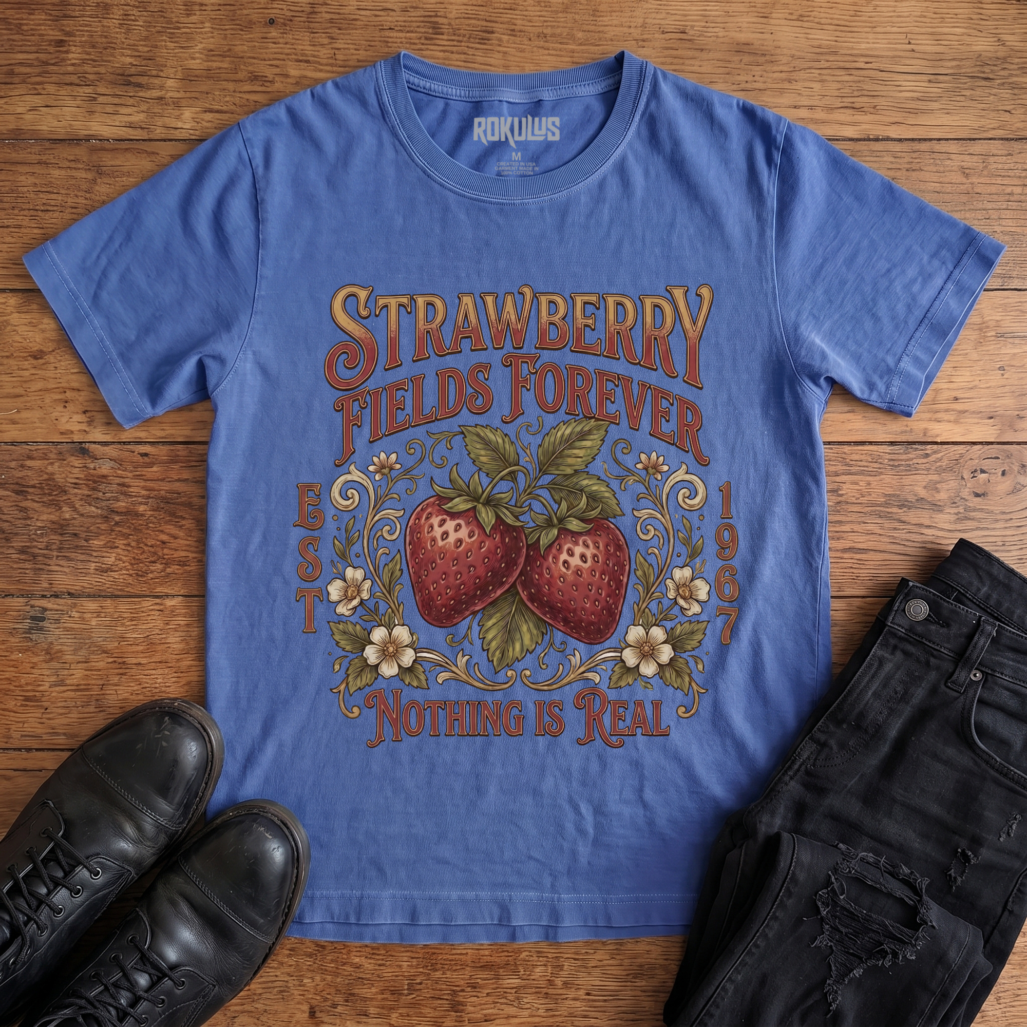 Washed royal blue Rokulus Strawberry Fields Botanical vintage graphic tee laid flat on wood with black boots and jeans, front print showing two ripe strawberries in Victorian scrollwork and wildflowers with "Strawberry Fields Forever Nothing Is Real" lettering