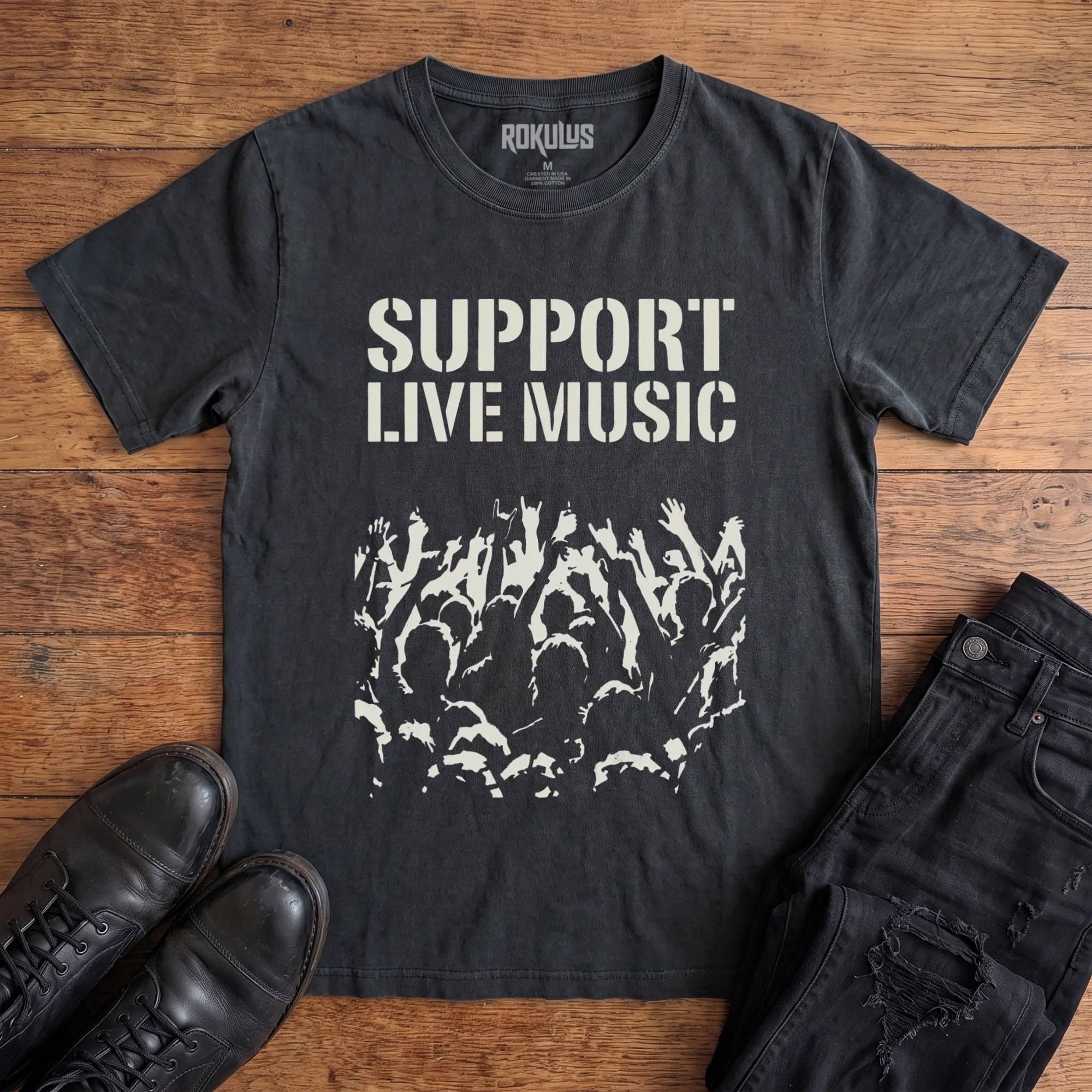 Washed black Rokulus Support Live Music vintage graphic tee laid flat on wood with black boots and jeans, front print showing "Support Live Music" in stencil type above a silhouetted concert crowd with raised hands