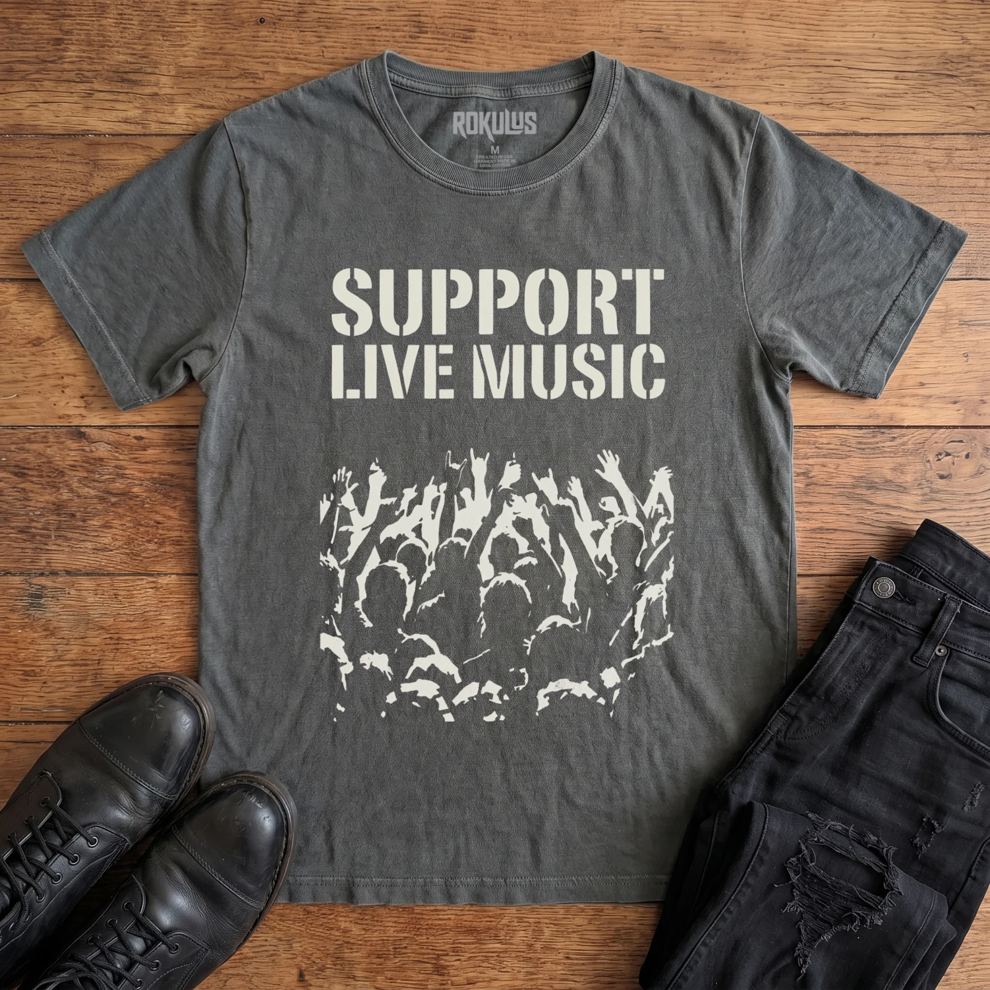 Washed charcoal Rokulus Support Live Music vintage graphic tee laid flat on wood with black boots and jeans, front print showing "Support Live Music" in stencil type above a silhouetted concert crowd with raised hands