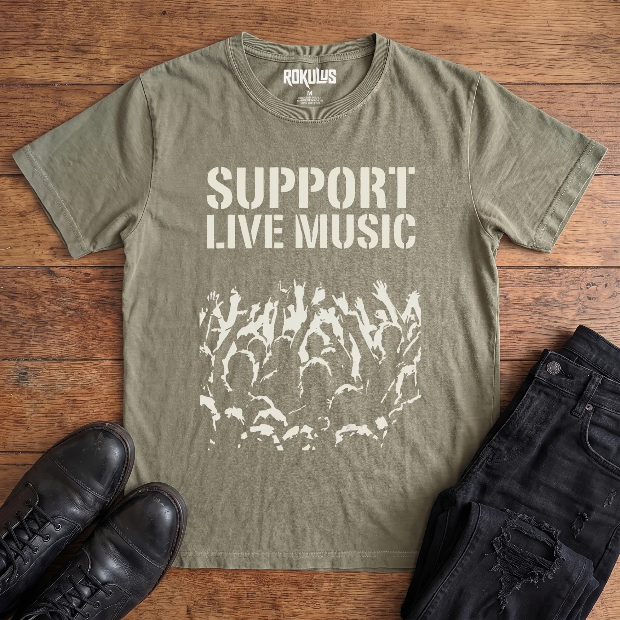 Washed olive Rokulus Support Live Music vintage graphic tee laid flat on wood with black boots and jeans, front print showing "Support Live Music" in stencil type above a silhouetted concert crowd with raised hands