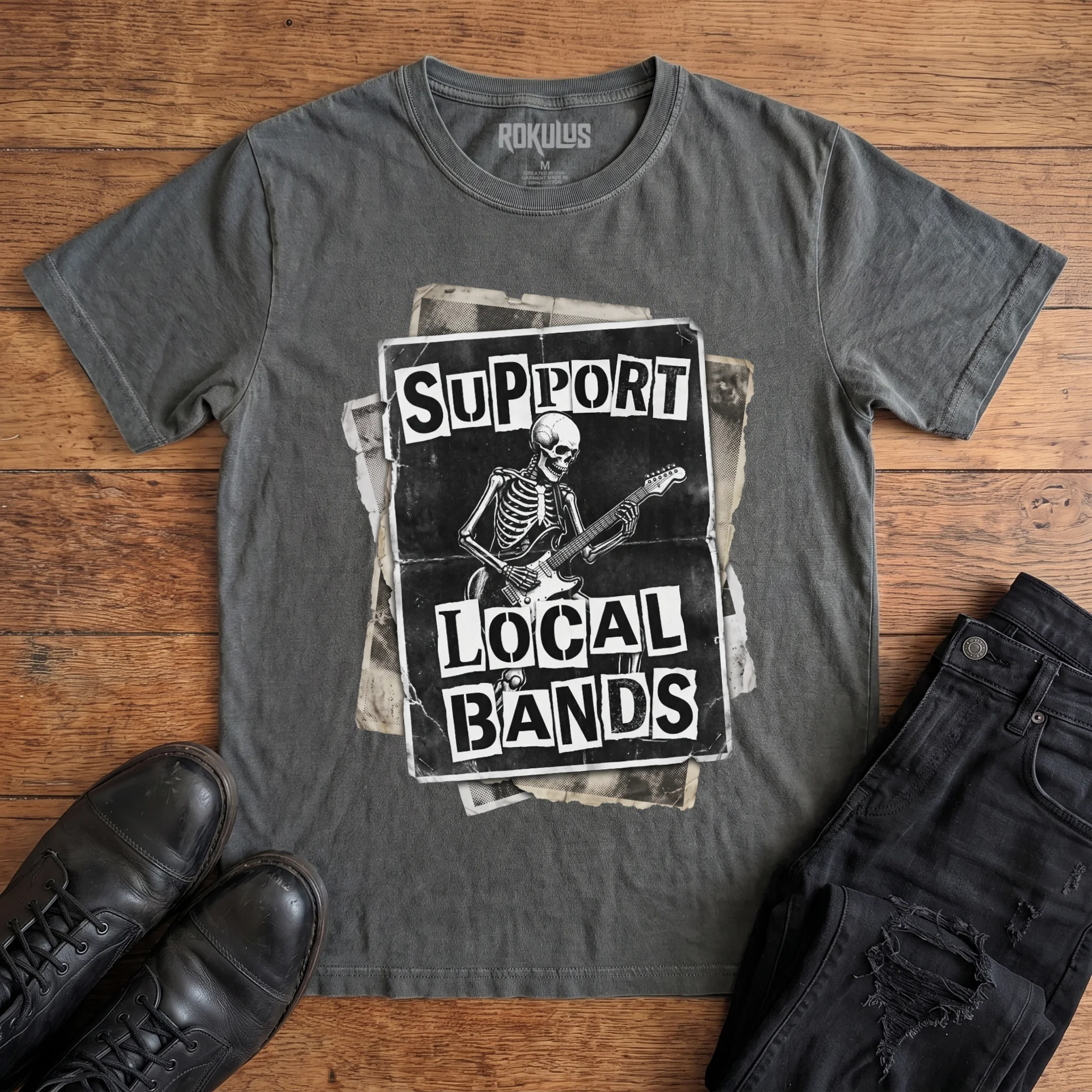 Washed charcoal Rokulus Support Local Bands vintage graphic tee laid flat on wood with black boots and jeans, front print showing a torn punk flyer with a skeleton playing electric guitar and "Support Local Bands" in ransom-note lettering