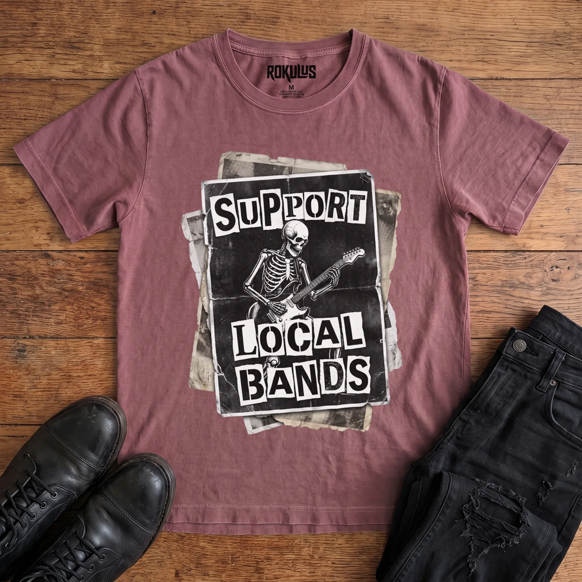 Washed maroon Rokulus Support Local Bands vintage graphic tee laid flat on wood with black boots and jeans, front print showing a torn punk flyer with a skeleton playing electric guitar and "Support Local Bands" in ransom-note lettering
