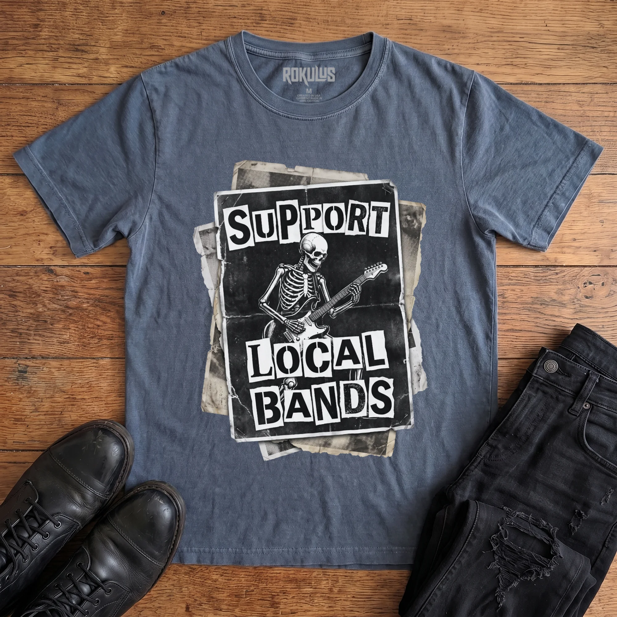Washed navy Rokulus Support Local Bands vintage graphic tee laid flat on wood with black boots and jeans, front print showing a torn punk flyer with a skeleton playing electric guitar and "Support Local Bands" in ransom-note lettering