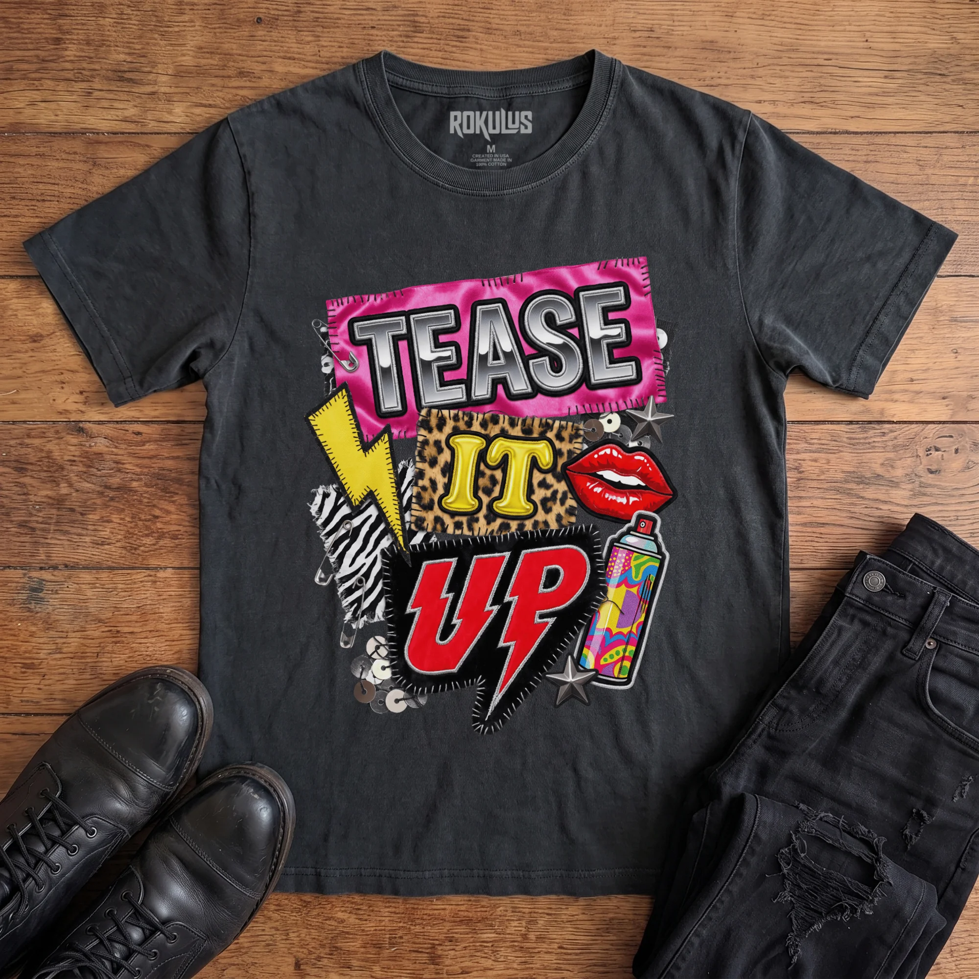 Washed black Rokulus Tease It Up vintage graphic tee laid flat on wood with black boots and jeans, front print showing an 80s glam collage with leopard and zebra patches, red lips, hairspray, and lightning bolts under chrome “Tease It Up” lettering