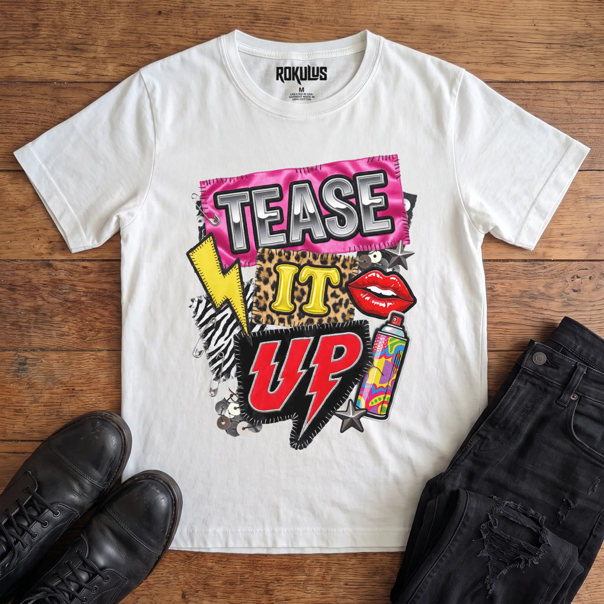 White Rokulus Tease It Up vintage graphic tee laid flat on wood with black boots and jeans, front print showing an 80s glam collage with leopard and zebra patches, red lips, hairspray, and lightning bolts under chrome “Tease It Up” lettering