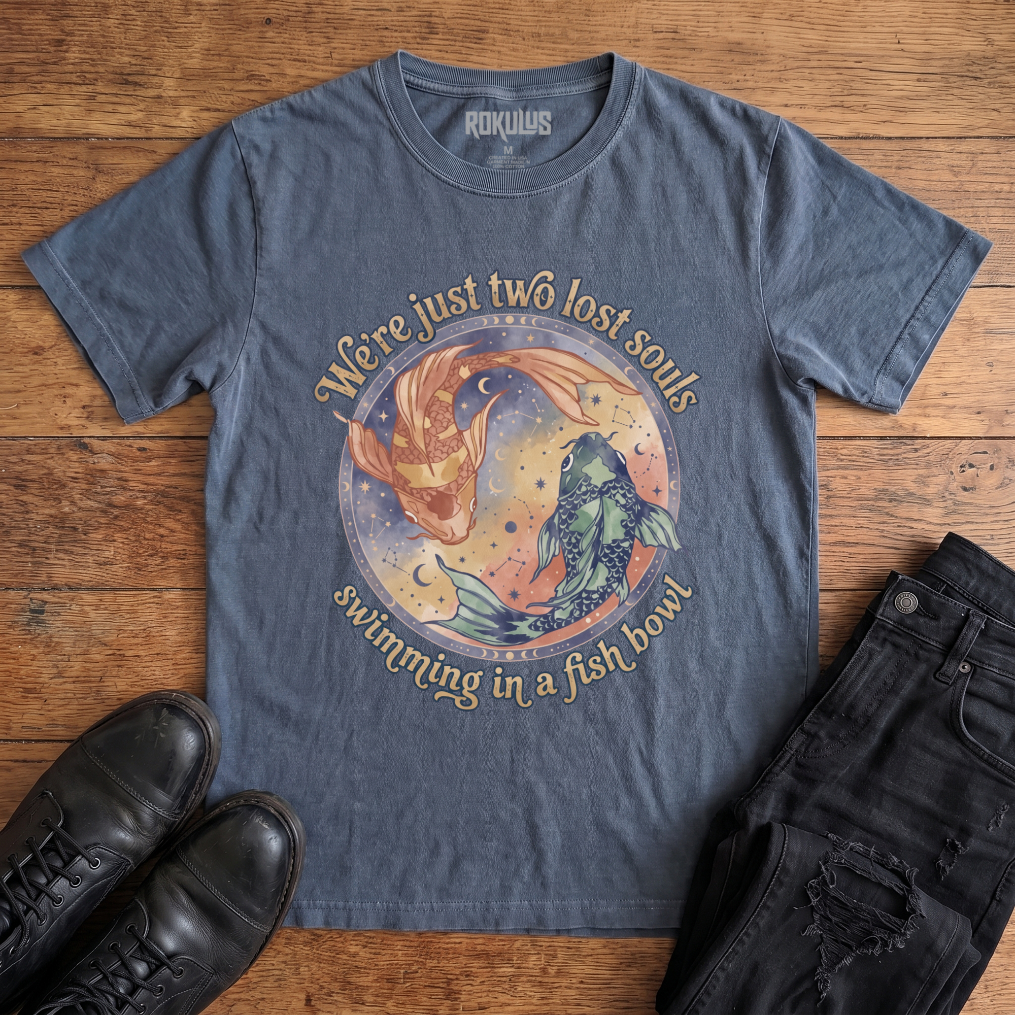 Washed navy blue Rokulus Two Lost Souls vintage graphic tee laid flat on wood with black boots and jeans, front print showing two watercolor koi in a celestial ring of moon phases and constellations with "We're just two lost souls swimming in a fish bowl" in retro script