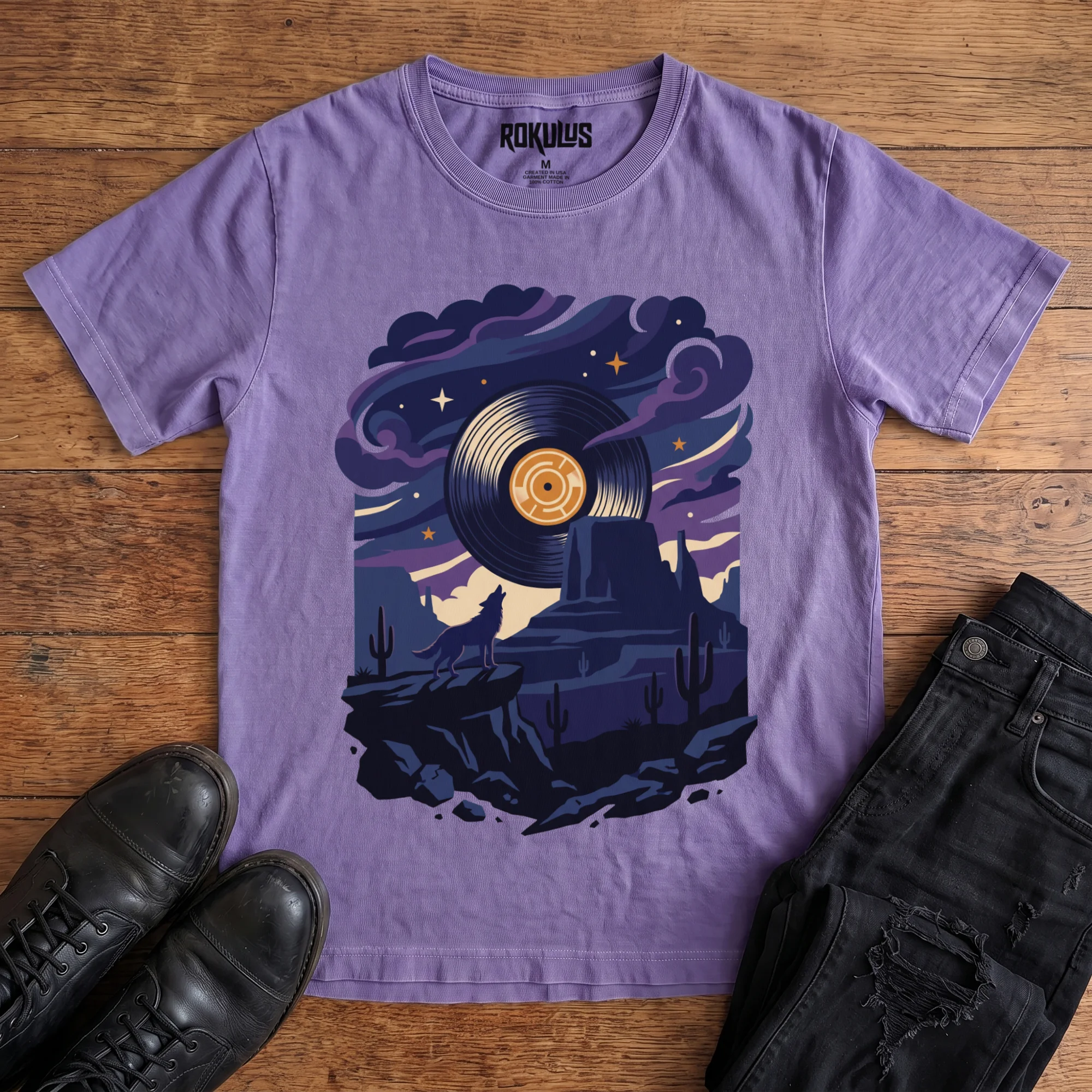 Washed purple Rokulus Vinyl Coyote vintage graphic tee laid flat on wood with black boots and jeans, front print showing a coyote howling at a vinyl-record moon over desert mesas and cactus under a starlit purple sky