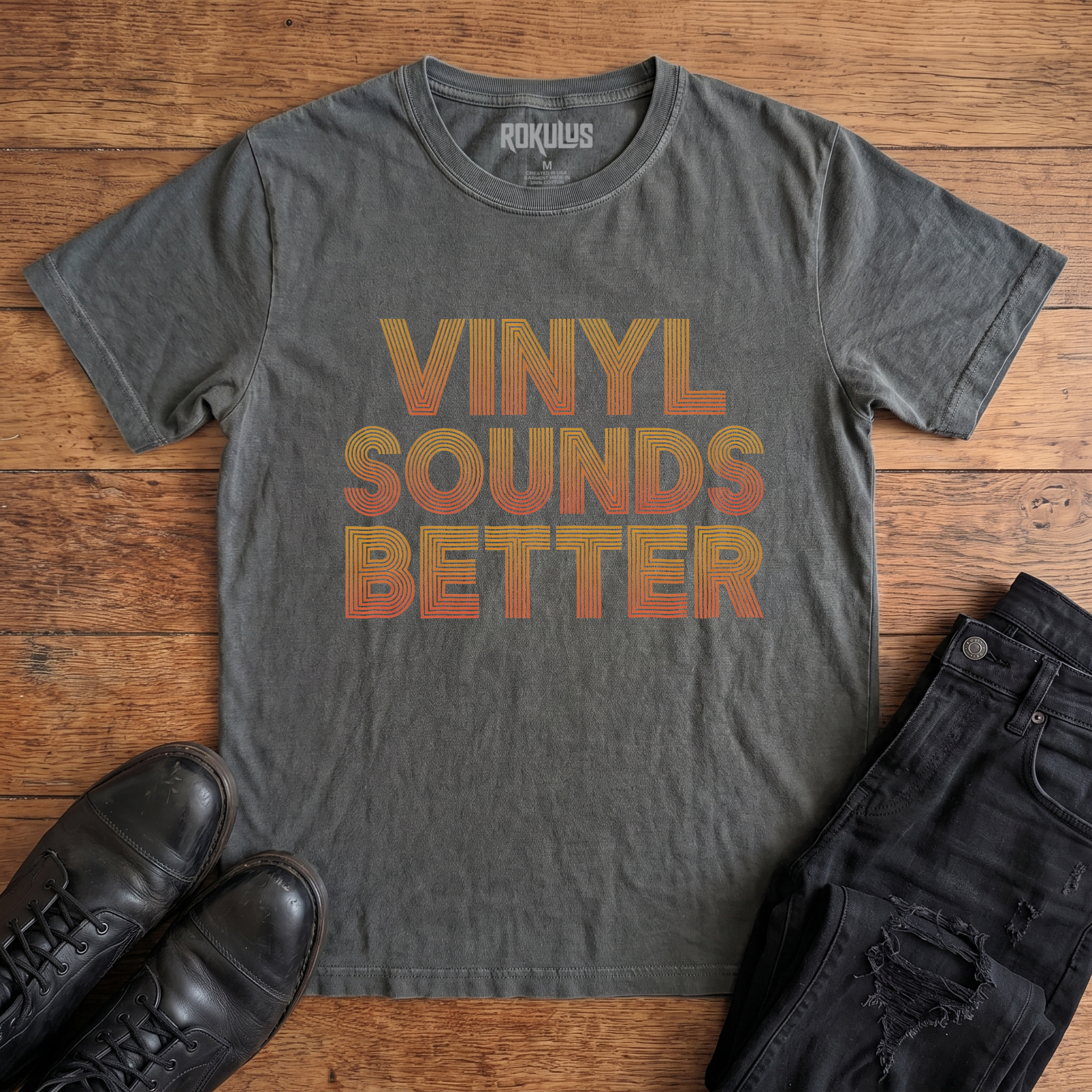 Washed charcoal Rokulus Vinyl Sounds Better vintage graphic tee laid flat on wood with black boots and jeans, front print showing "Vinyl Sounds Better" stacked in three lines of retro layered-line type in a golden-to-orange gradient