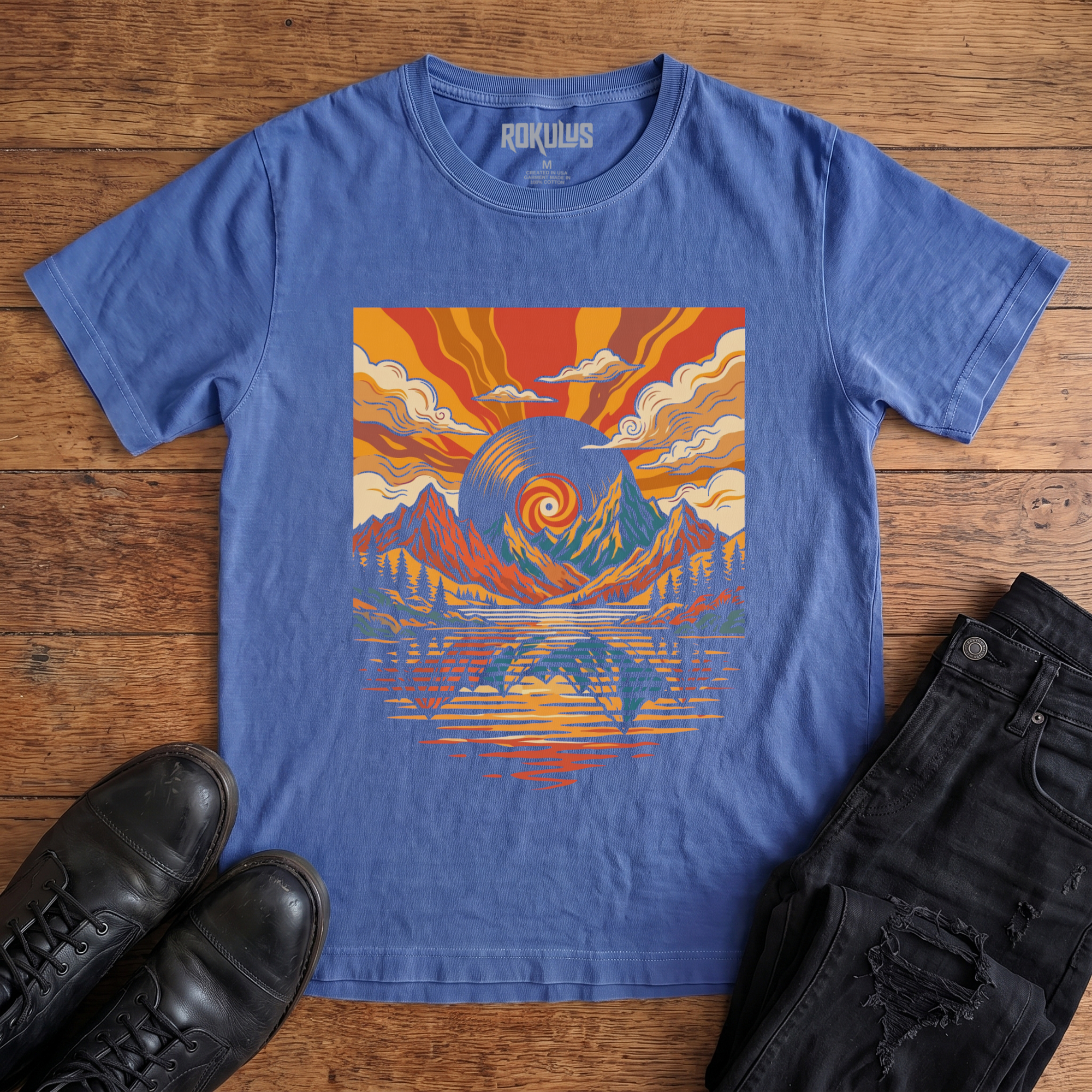 Washed royal blue Rokulus Vinyl Sunset vintage graphic tee laid flat on wood with black boots and jeans, front print showing a retro 70s landscape with a vinyl record sun over mountains, pines, and a reflecting lake in warm sunset tones