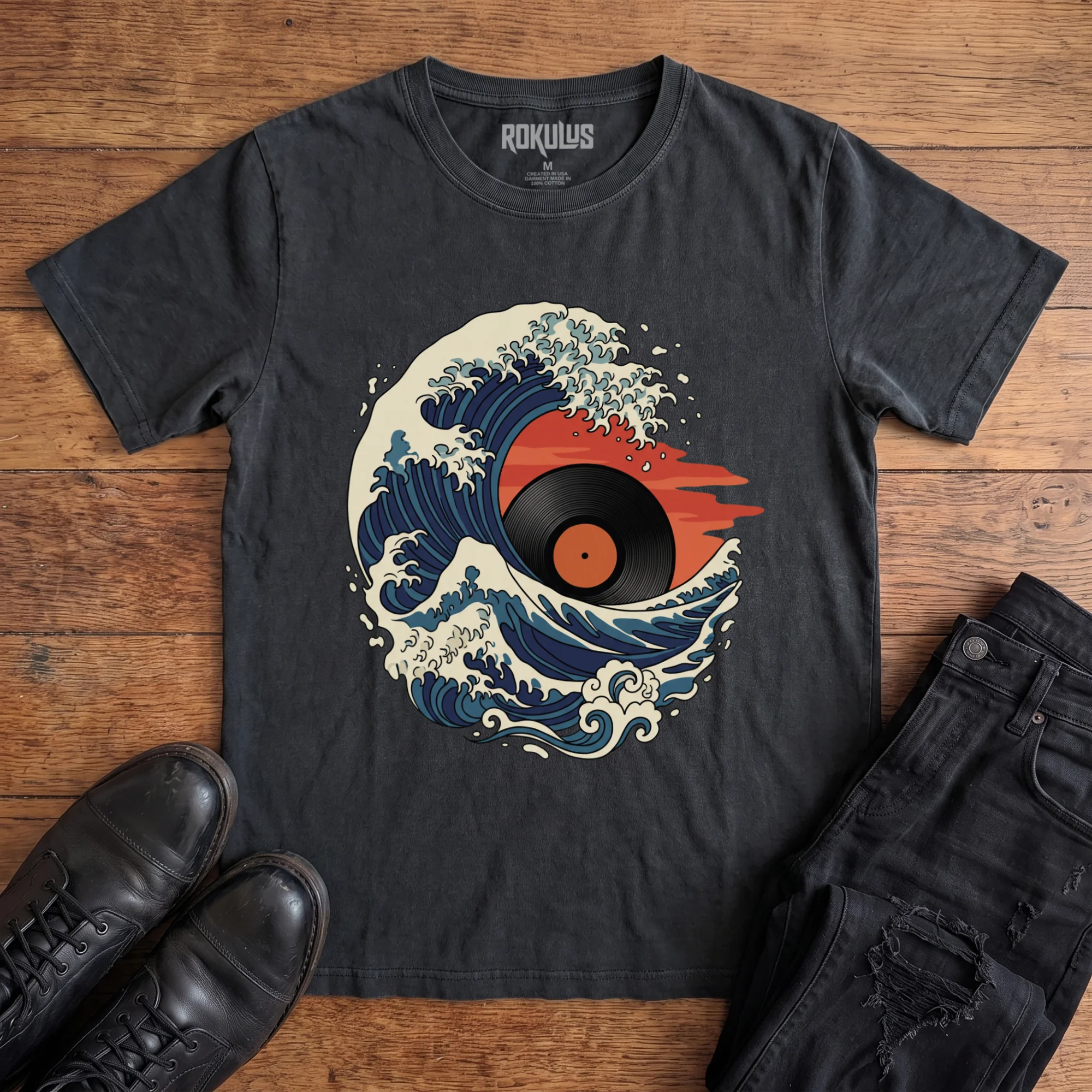 Washed black Rokulus Great Wave Vinyl vintage graphic tee laid flat on wood with black boots and jeans, front print showing a Hokusai-style wave curling around a vinyl record sun in indigo, cream, and burnt orange