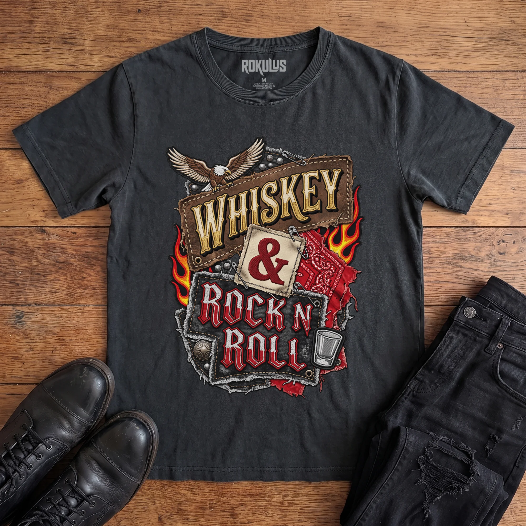 Washed black Rokulus Whiskey & Rock N Roll vintage graphic tee laid flat on wood with black boots and jeans, front print showing an embroidered biker-patch collage with a bald eagle, flames, red bandana, studs, and a shot glass around stitched “Whiskey & Rock N Roll” lettering