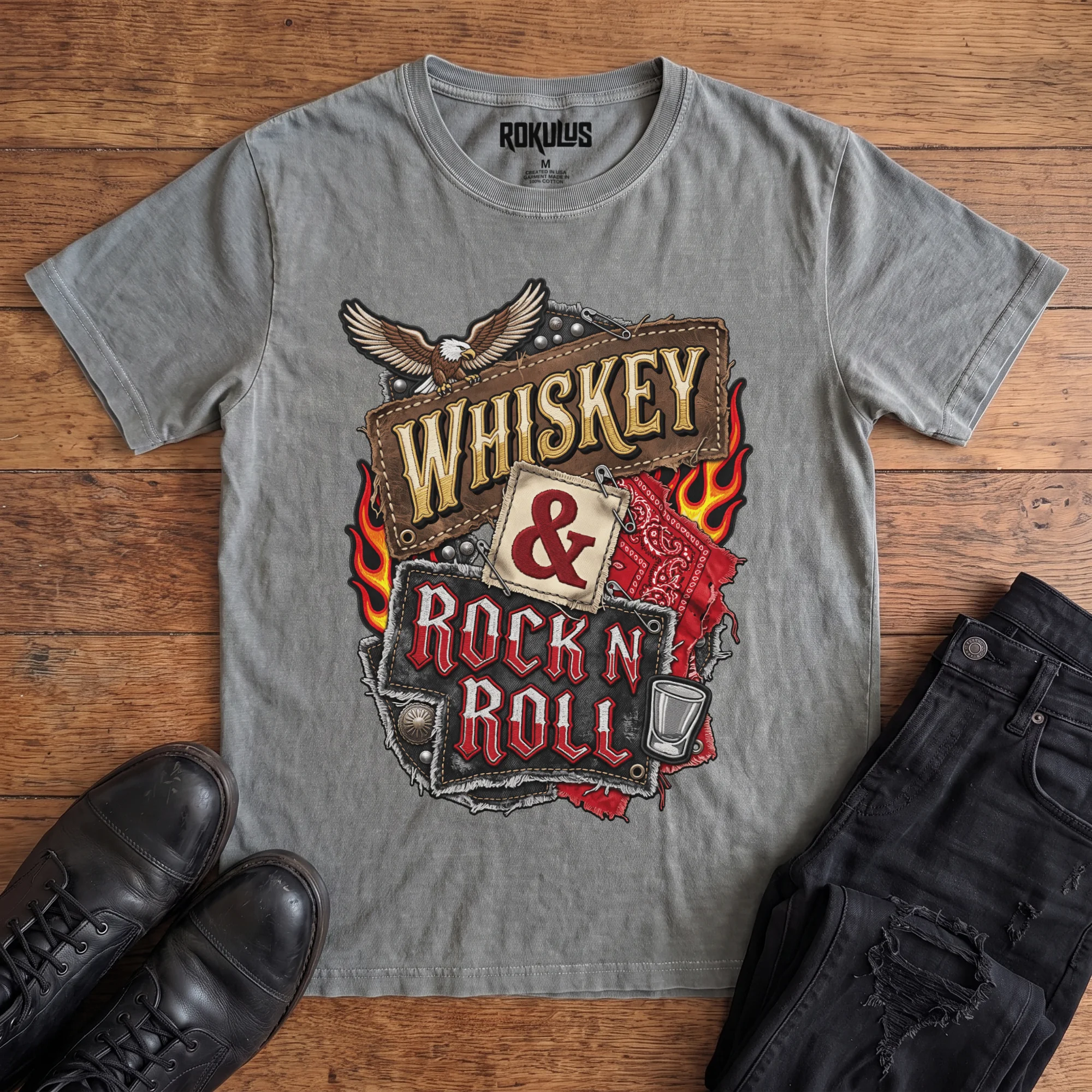 Washed grey Rokulus Whiskey & Rock N Roll vintage graphic tee laid flat on wood with black boots and jeans, front print showing an embroidered biker-patch collage with a bald eagle, flames, red bandana, studs, and a shot glass around stitched “Whiskey & Rock N Roll” lettering