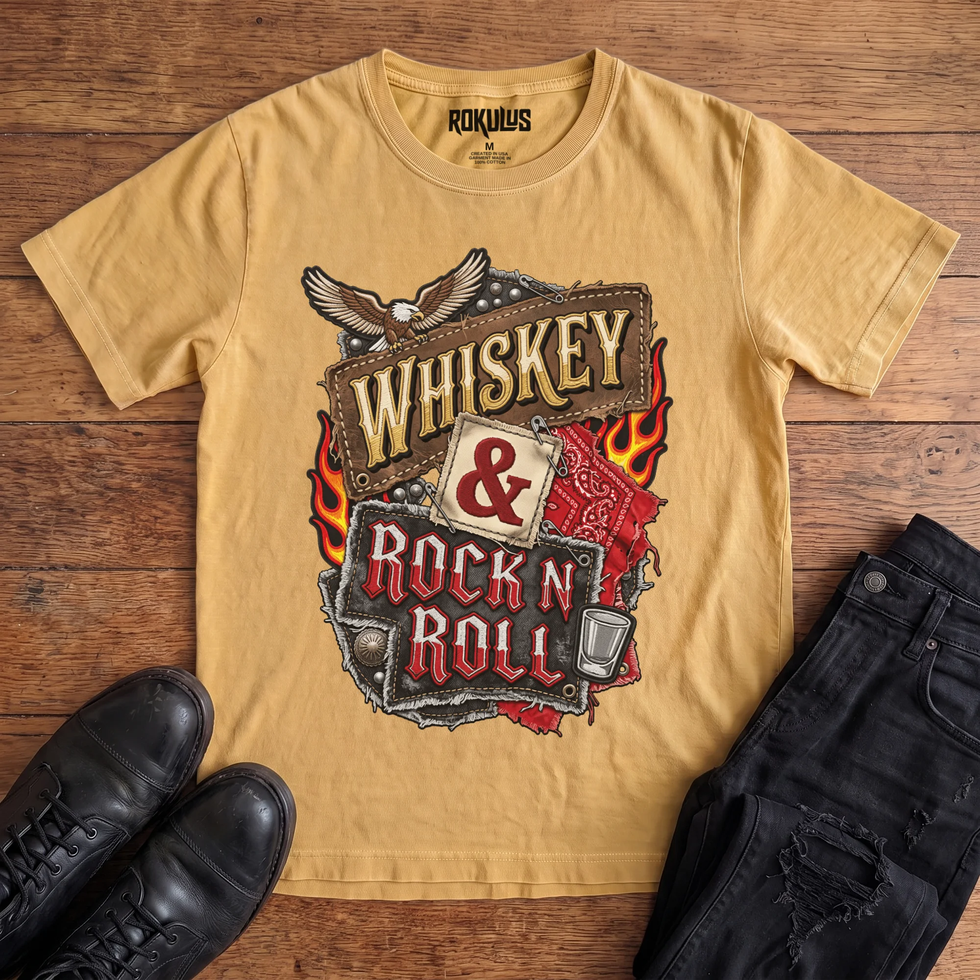 Washed mustard Rokulus Whiskey & Rock N Roll vintage graphic tee laid flat on wood with black boots and jeans, front print showing an embroidered biker-patch collage with a bald eagle, flames, red bandana, studs, and a shot glass around stitched “Whiskey & Rock N Roll” lettering