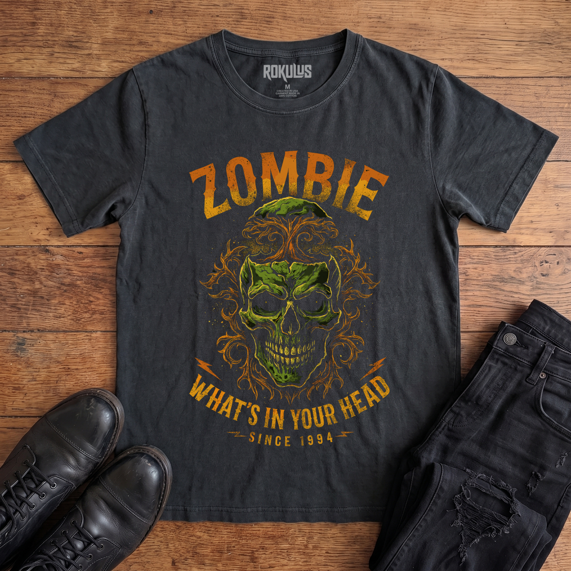 Washed black Rokulus Zombie vintage graphic tee laid flat on wood with black boots and jeans, front print showing a decaying green skull wreathed in orange filigree under arched "Zombie" lettering and a "What's In Your Head Since 1994" banner